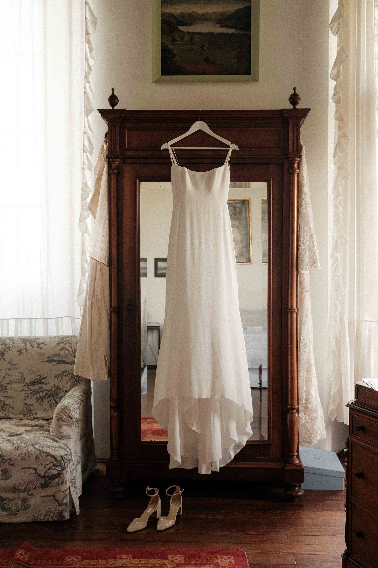 A white dress hangs on a wooden wardrobe in a sunlit bedroom with hardwood floors and sheer curtains.