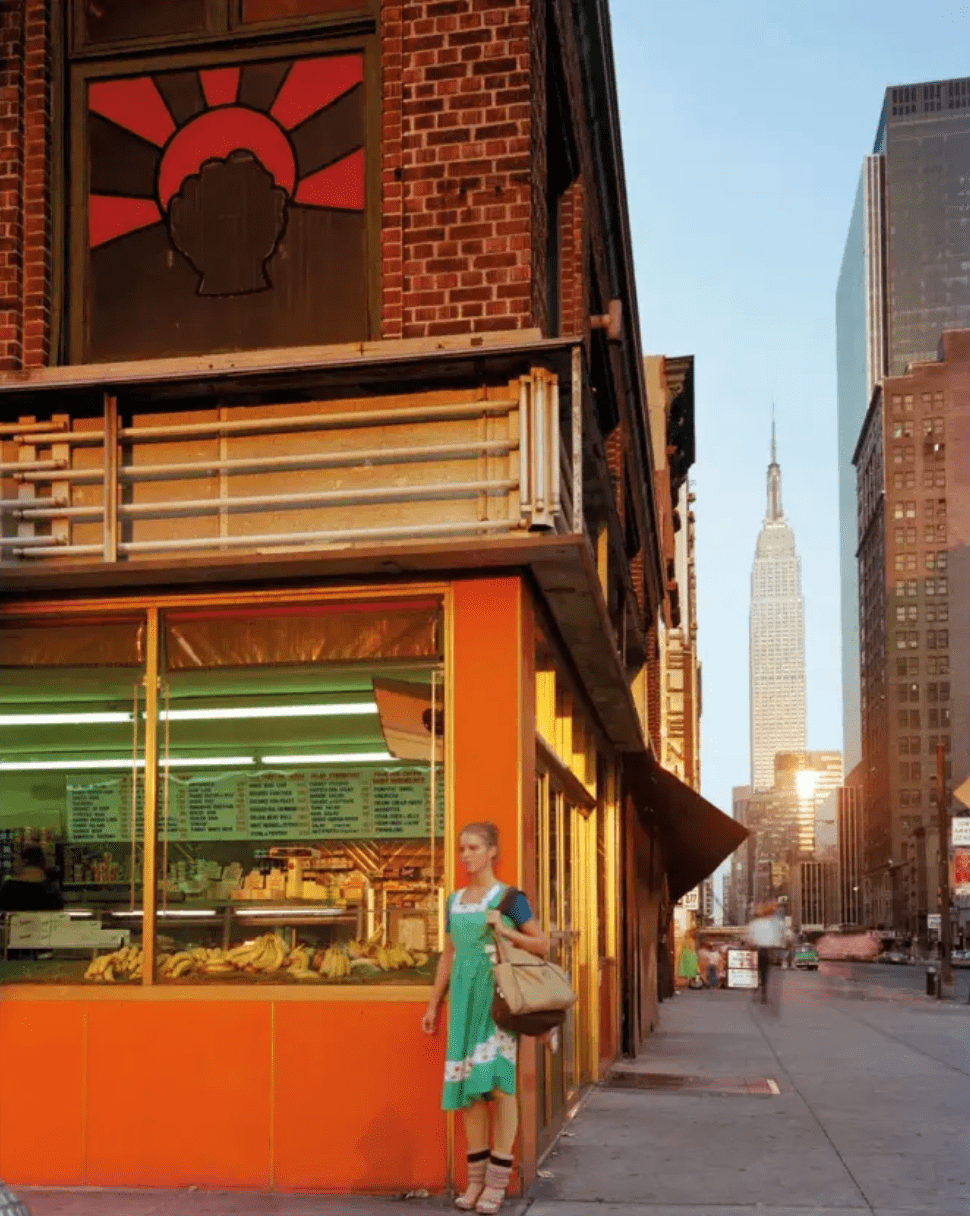 A figure in a green dress stands against orange-lit storefront windows on a brick building at sunset.