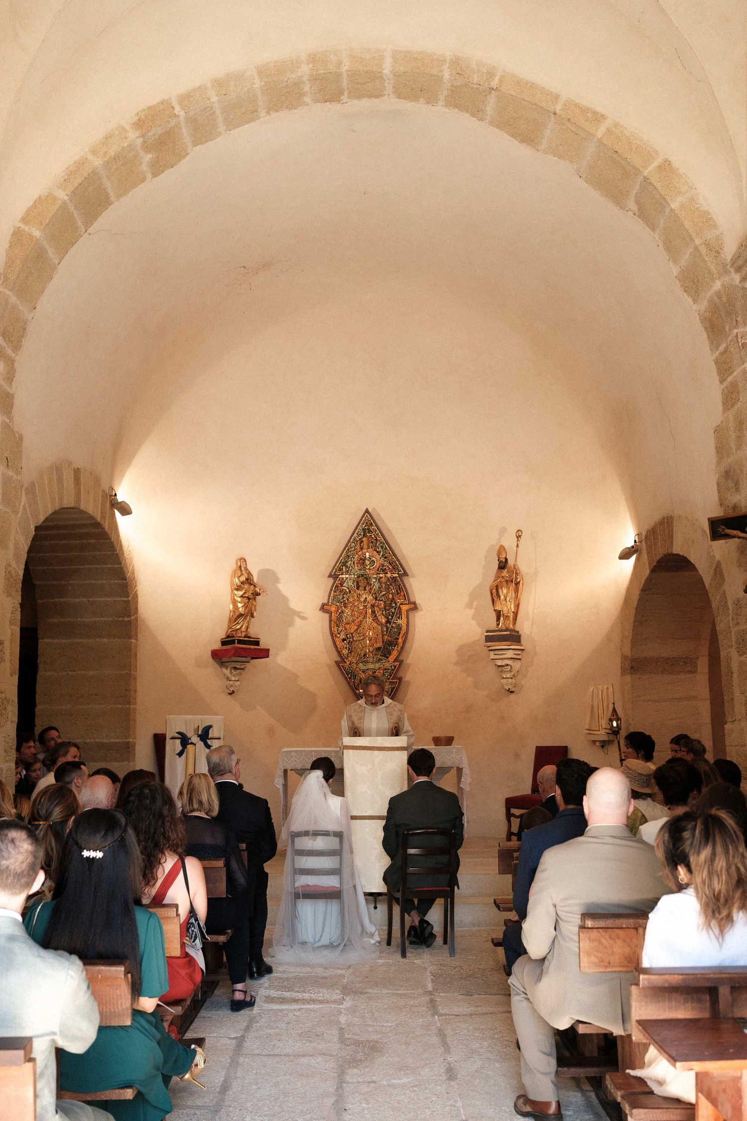 People gathered in a small chapel with stone arches and religious statues mounted on the white walls during a service.