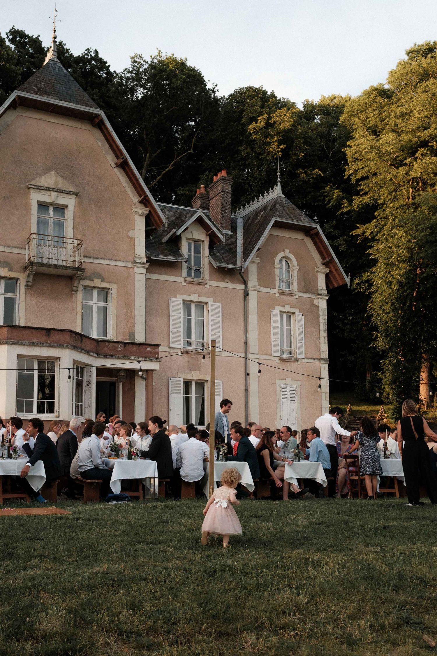 Elegant outdoor wedding reception at a vintage pink chateau with guests dining on the lawn at sunset.