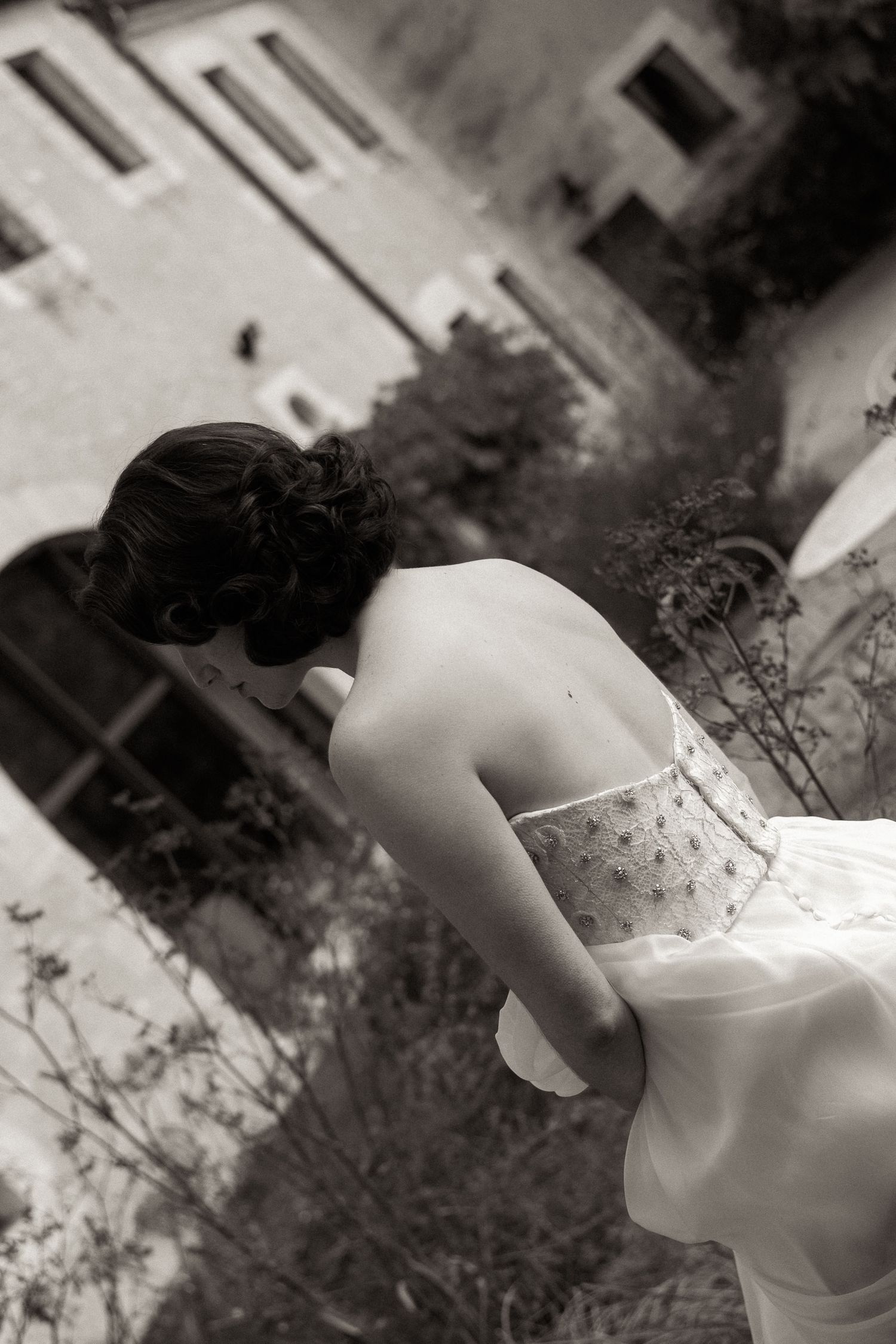 Black and white artistic photograph showing a person in a white dress leaning over a balcony, viewed from behind.