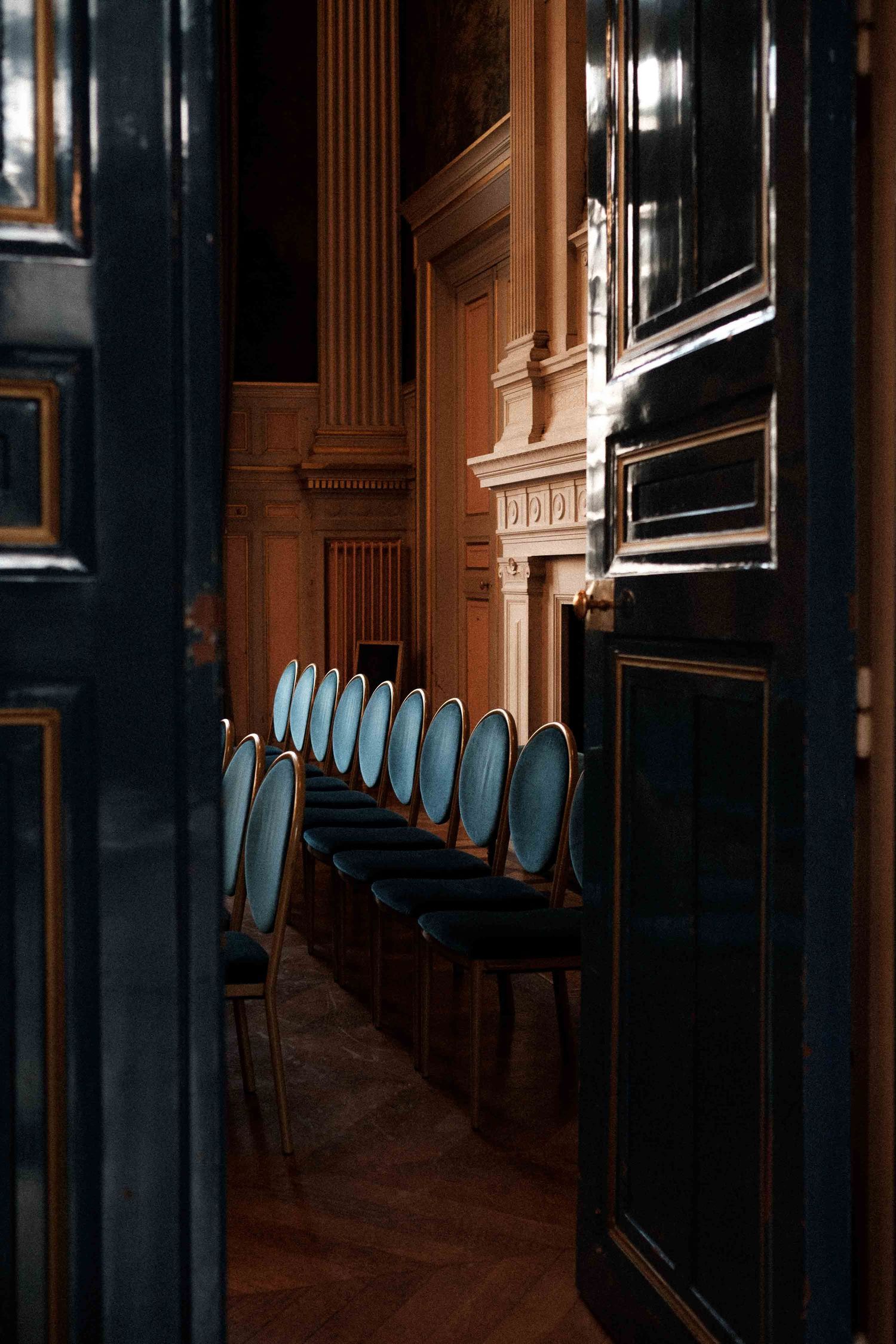 Row of blue chairs line a dark wood-paneled hallway with ornate architectural details in warm lighting.