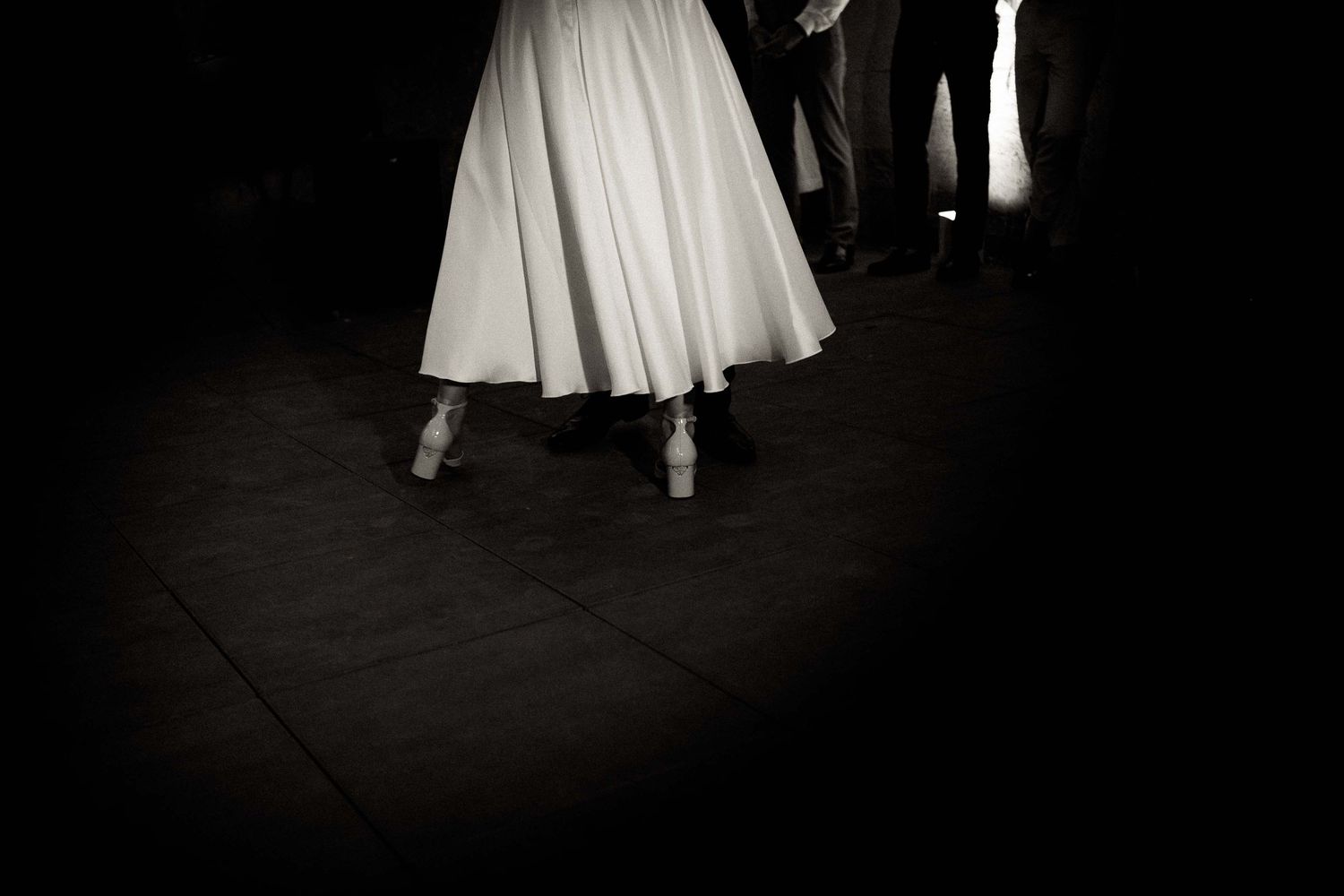 A black and white photograph shows a flowing white skirt and dance shoes on a dark floor during a performance.