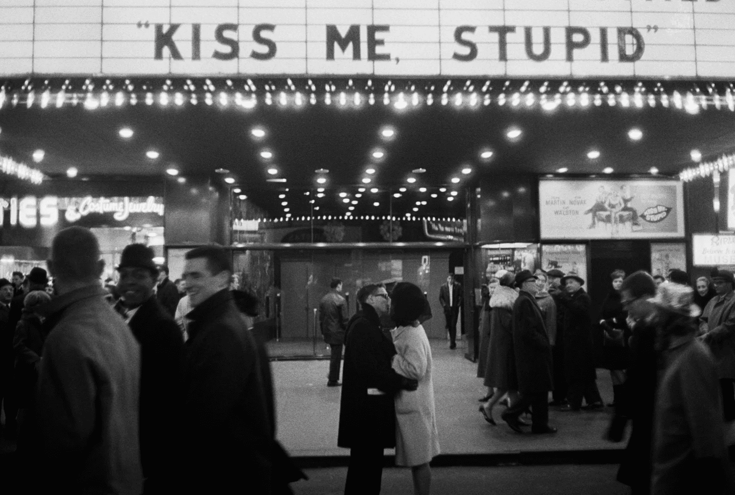 Black and white photo of a neon sign that reads 'KISS ME STUPID' glowing against a dark background.