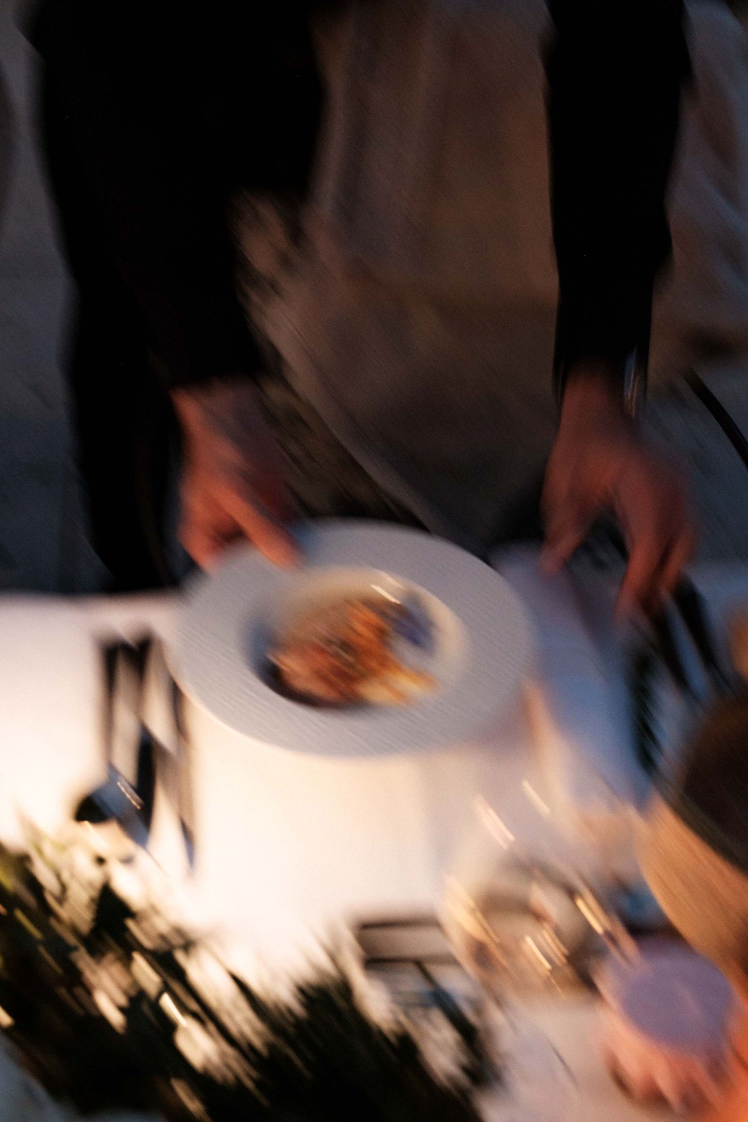 A blurred motion shot of hands serving food on a white plate in dim lighting.