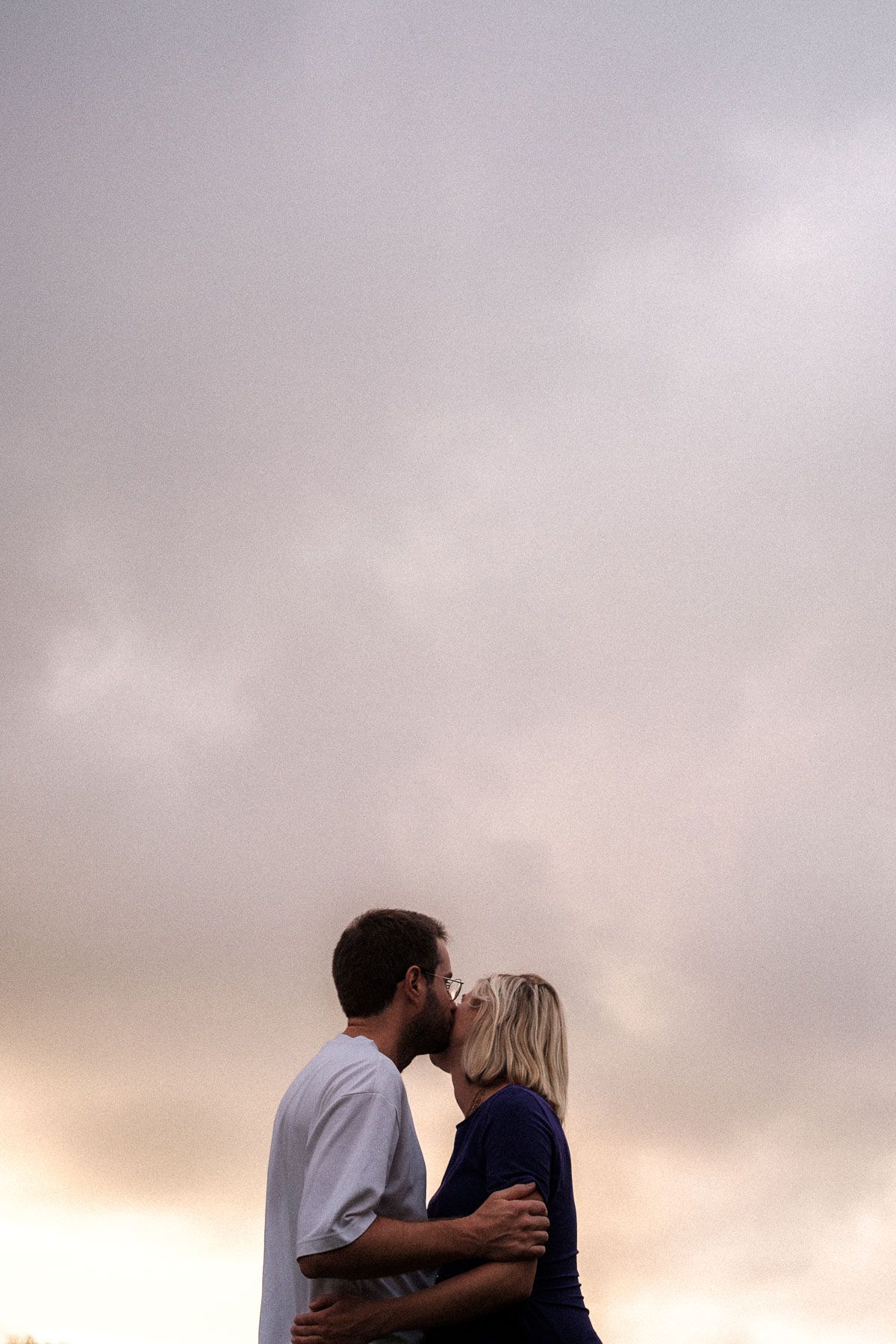 A couple sharing a romantic kiss at sunset with cloudy skies in the background.