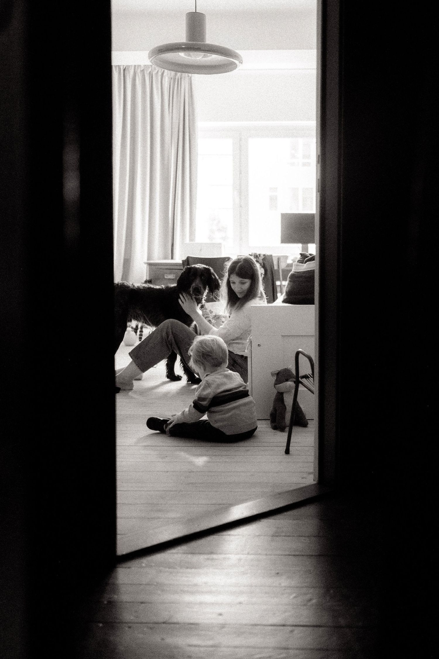 A black and white candid view through a doorway of a family gathering with a small child on the floor.