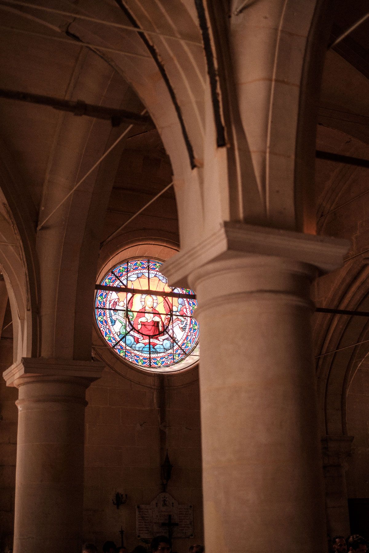 Ornate stained glass windows illuminate a vaulted stone ceiling with arched columns in a historic church interior.