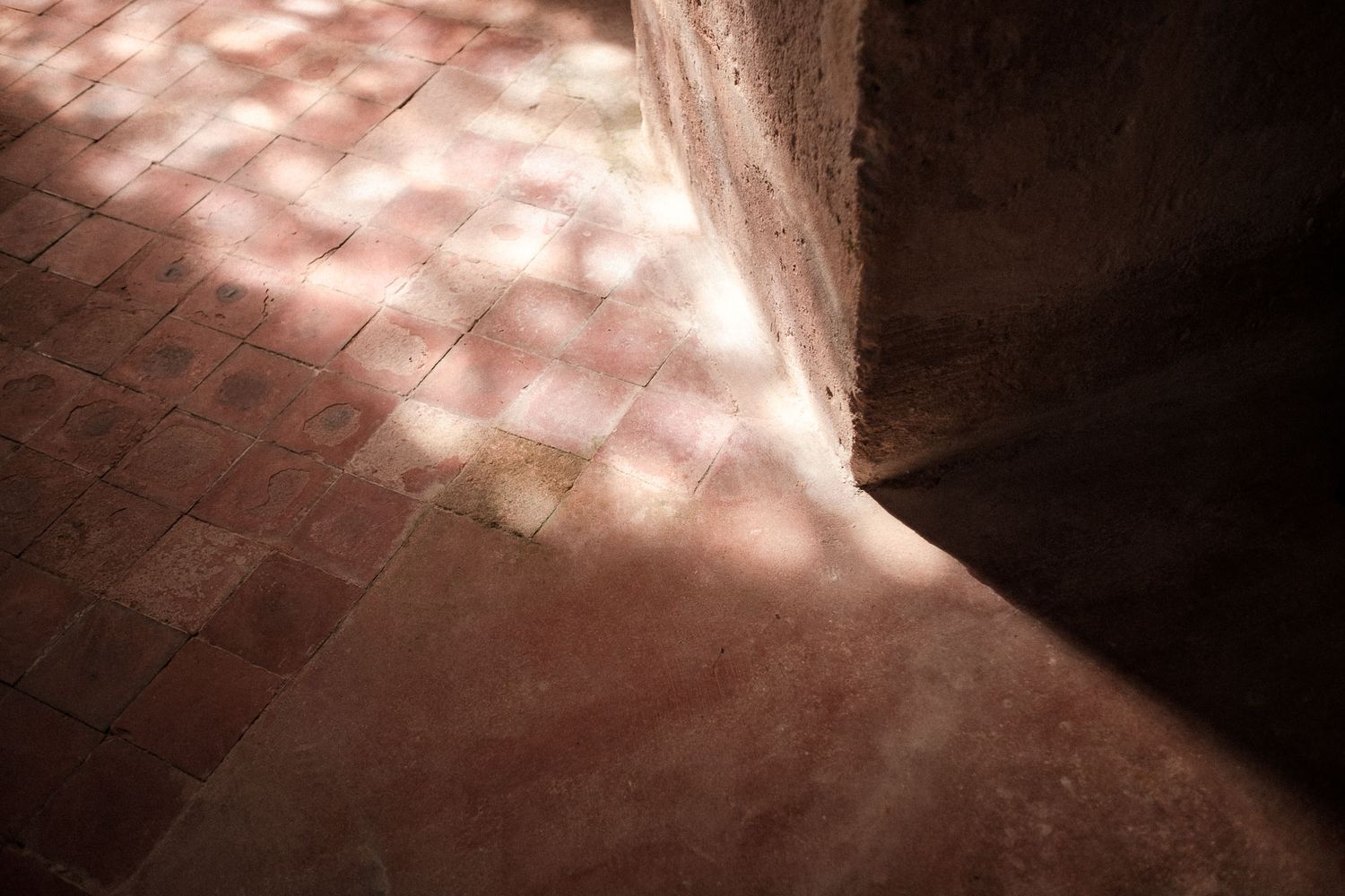 Sunlight casts dappled shadows on red brick floor near a curved stone wall in a dimly lit space.