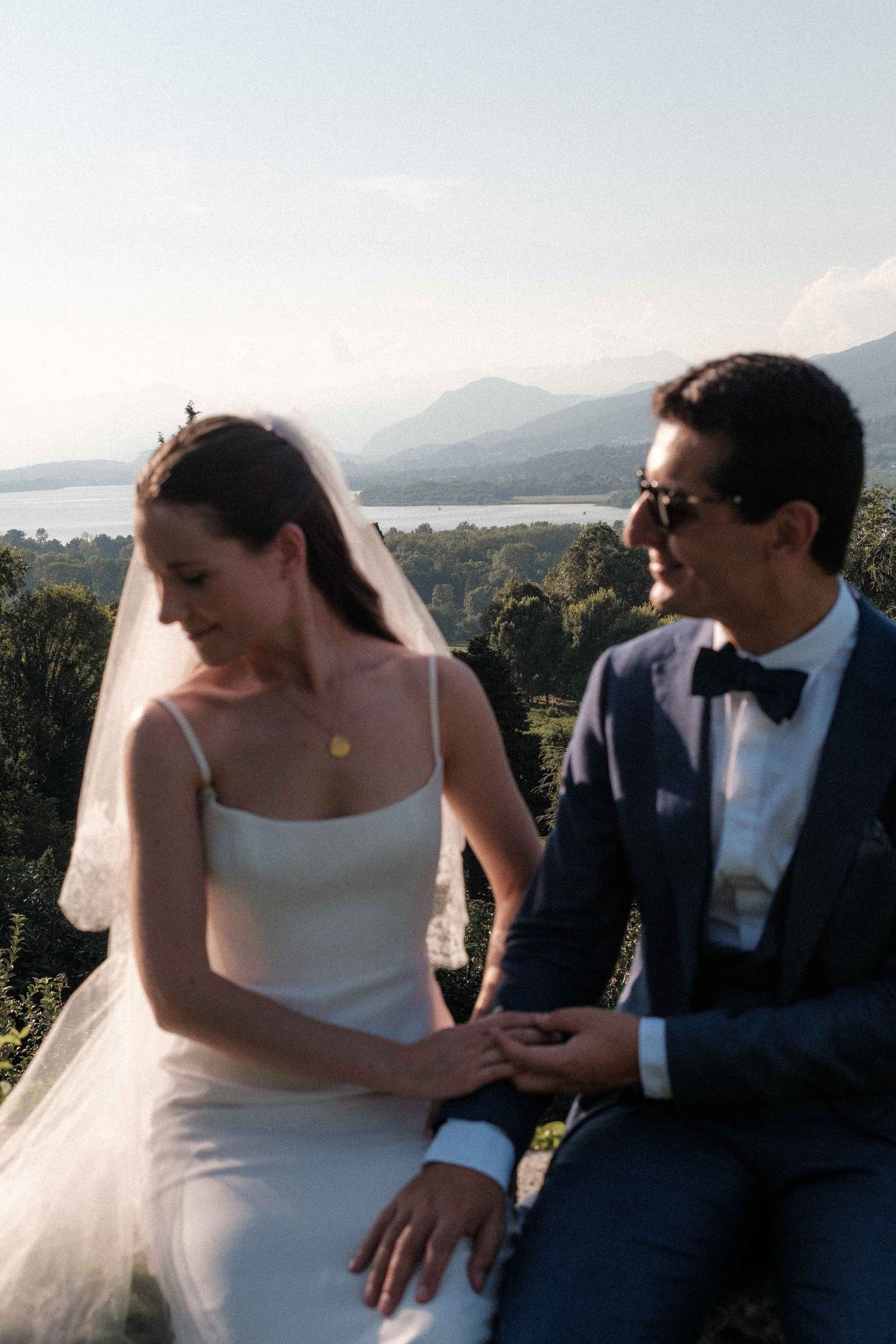 A couple in wedding attire sits together against a scenic mountain and lake backdrop on their special day.