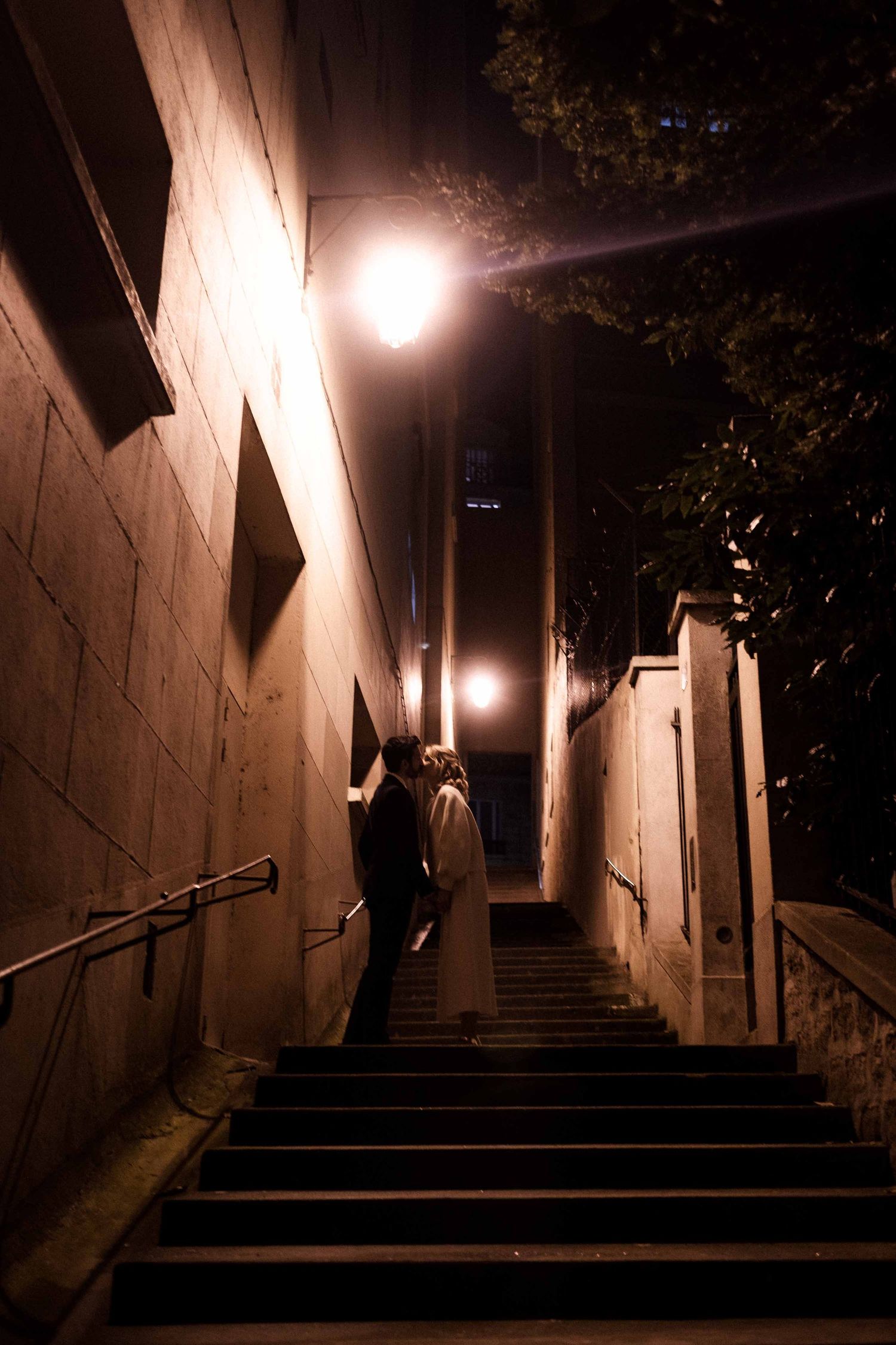 A dimly lit concrete stairway at night with handrails and dramatic shadows cast by overhead lighting.