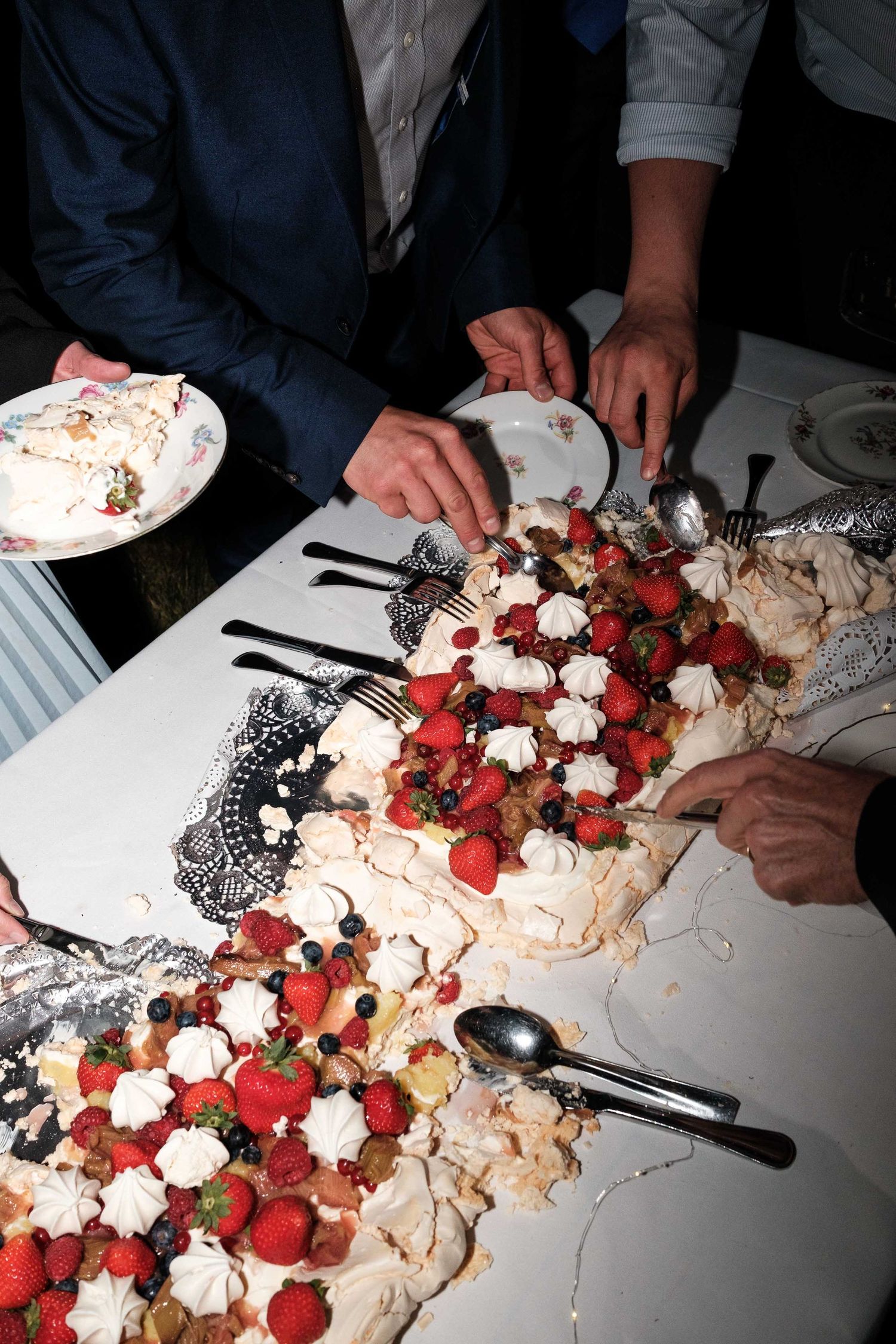 A dessert table shows hands assembling a pavlova with fresh berries and whipped cream toppings.