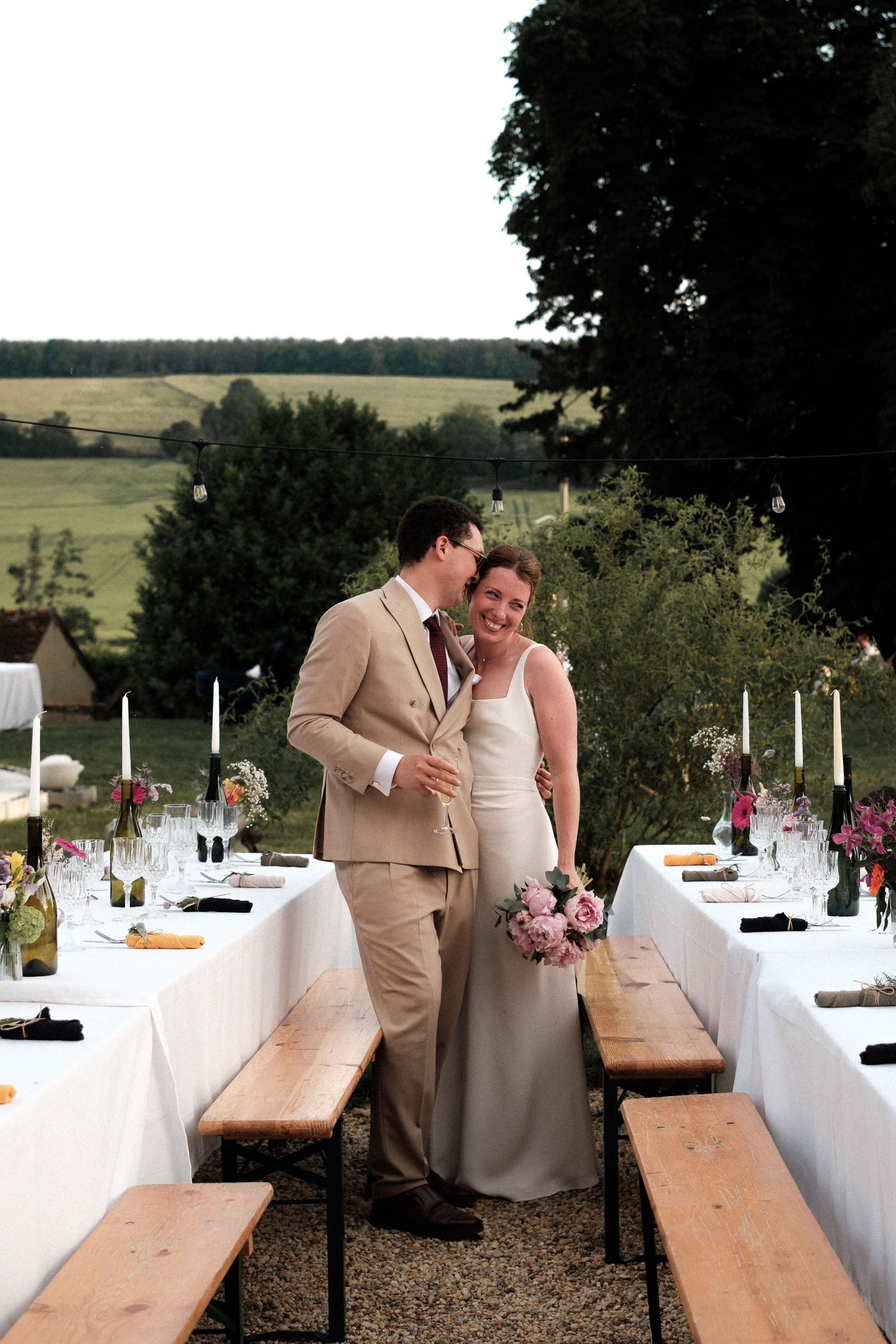 A couple shares a tender moment at an outdoor wedding reception with long wooden tables and white linens.