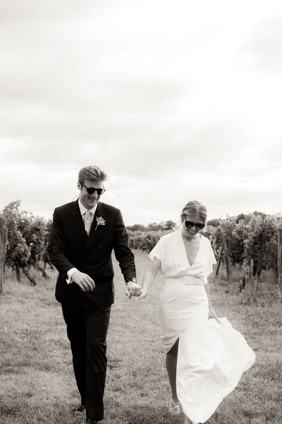 A bride and groom in sunglasses walk together through a vineyard on their wedding day, captured in black and white.