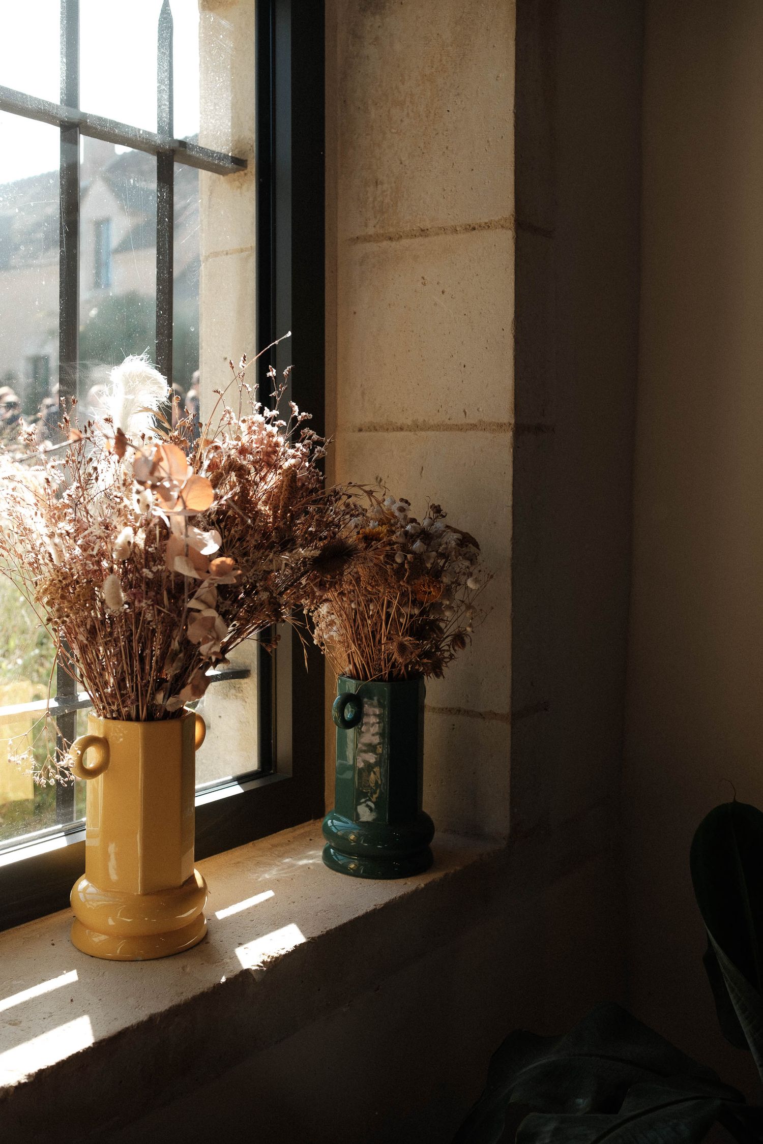 Dried flowers in yellow ceramic vases line a windowsill, casting dramatic shadows in natural sunlight.
