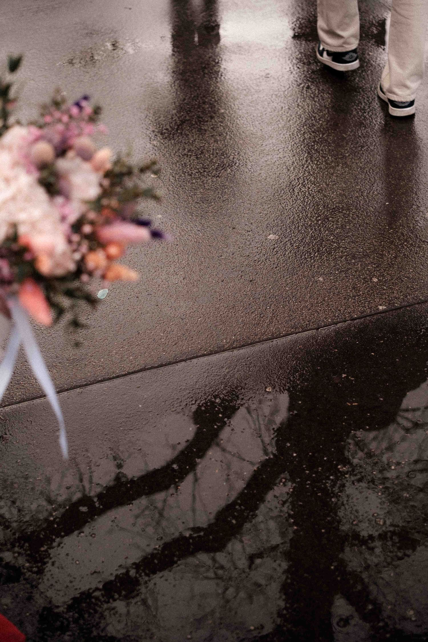 A series of reflections in wet pavement showing legs walking and pink floral bouquets creating a moody urban scene.