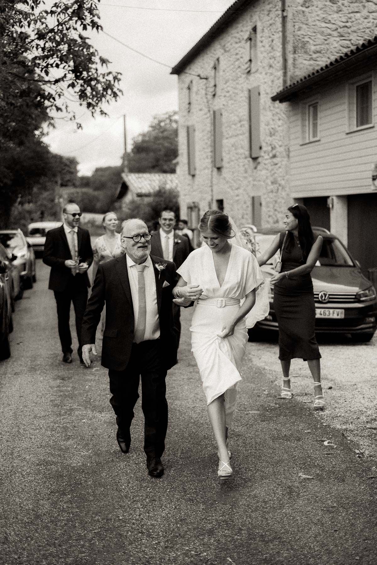 A black and white wedding photo showing guests walking along a rustic country road beside a stone building.
