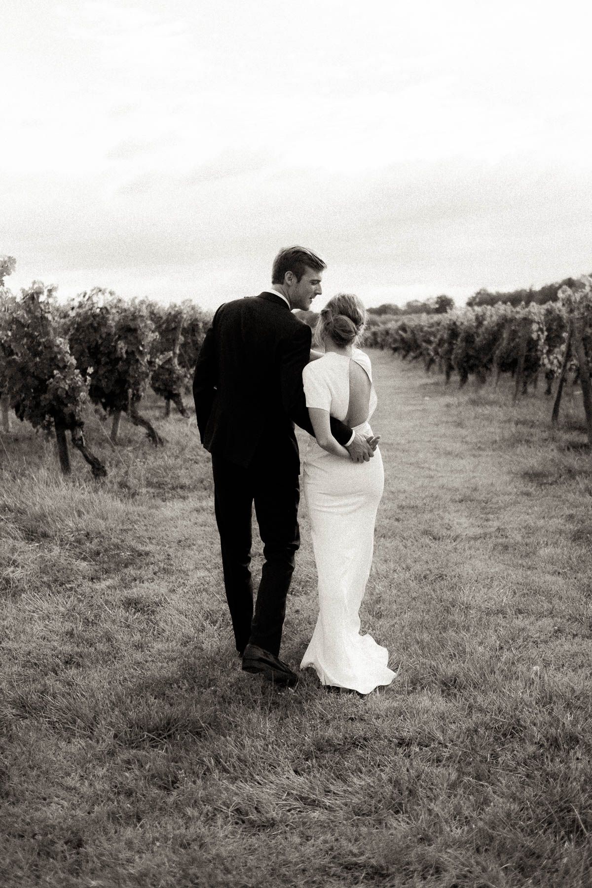 A couple in wedding attire walks through a vineyard in black and white photography.