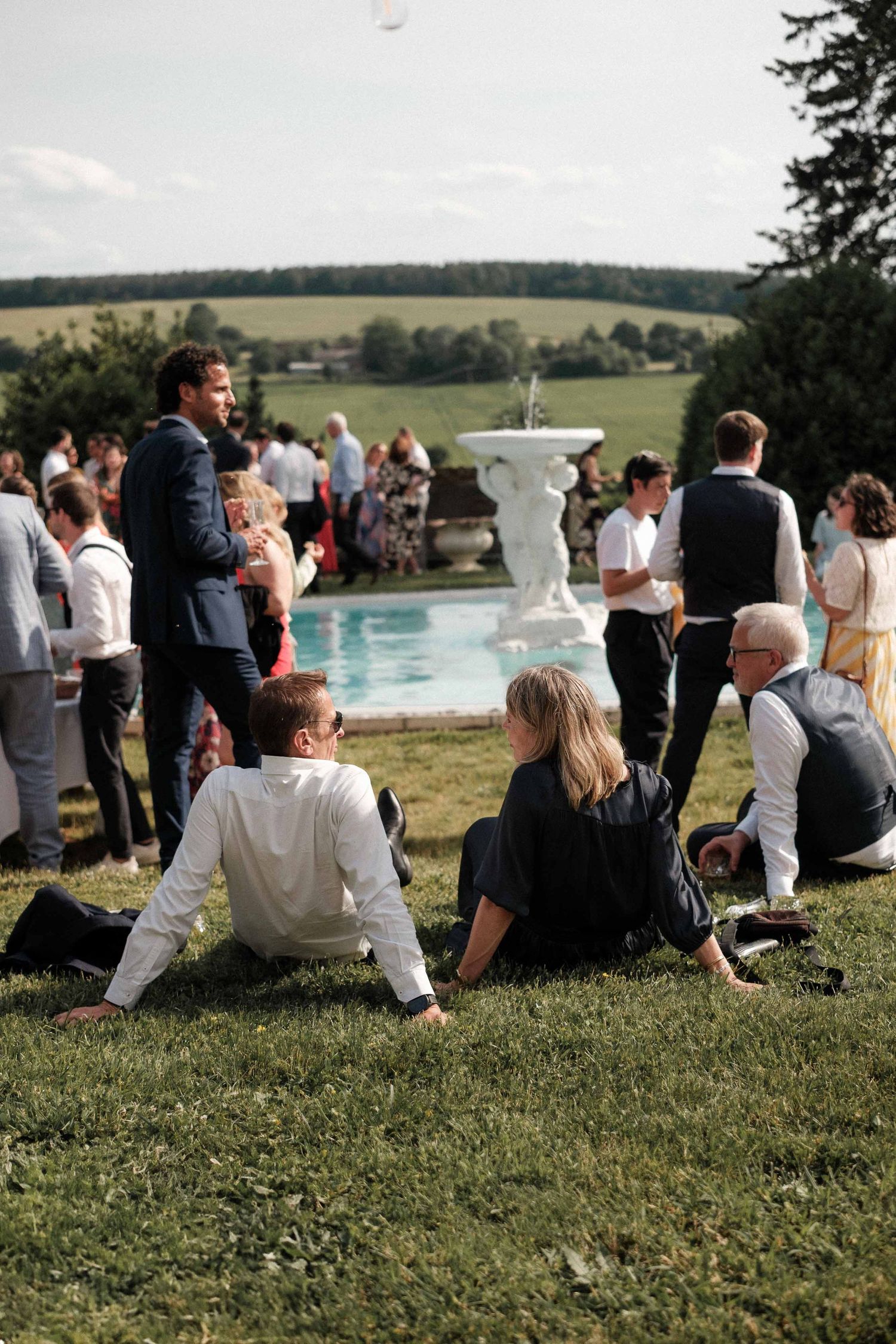 Wedding guests relax on a grassy hillside overlooking a scenic countryside view during an outdoor reception.
