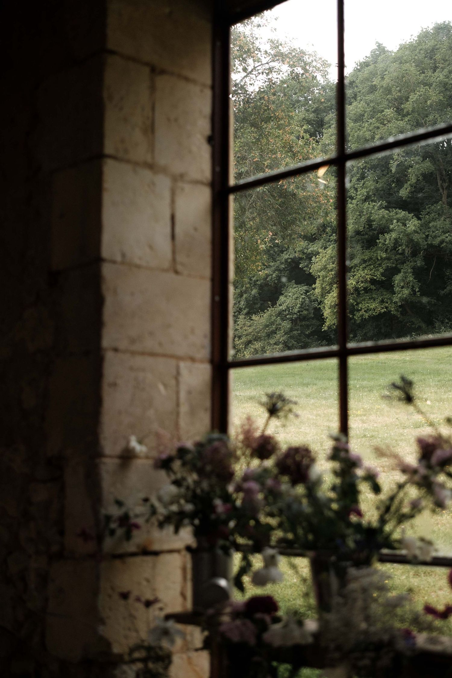 Rustic window view of a green meadow with trees in the background, framed by stone walls and delicate flowers.