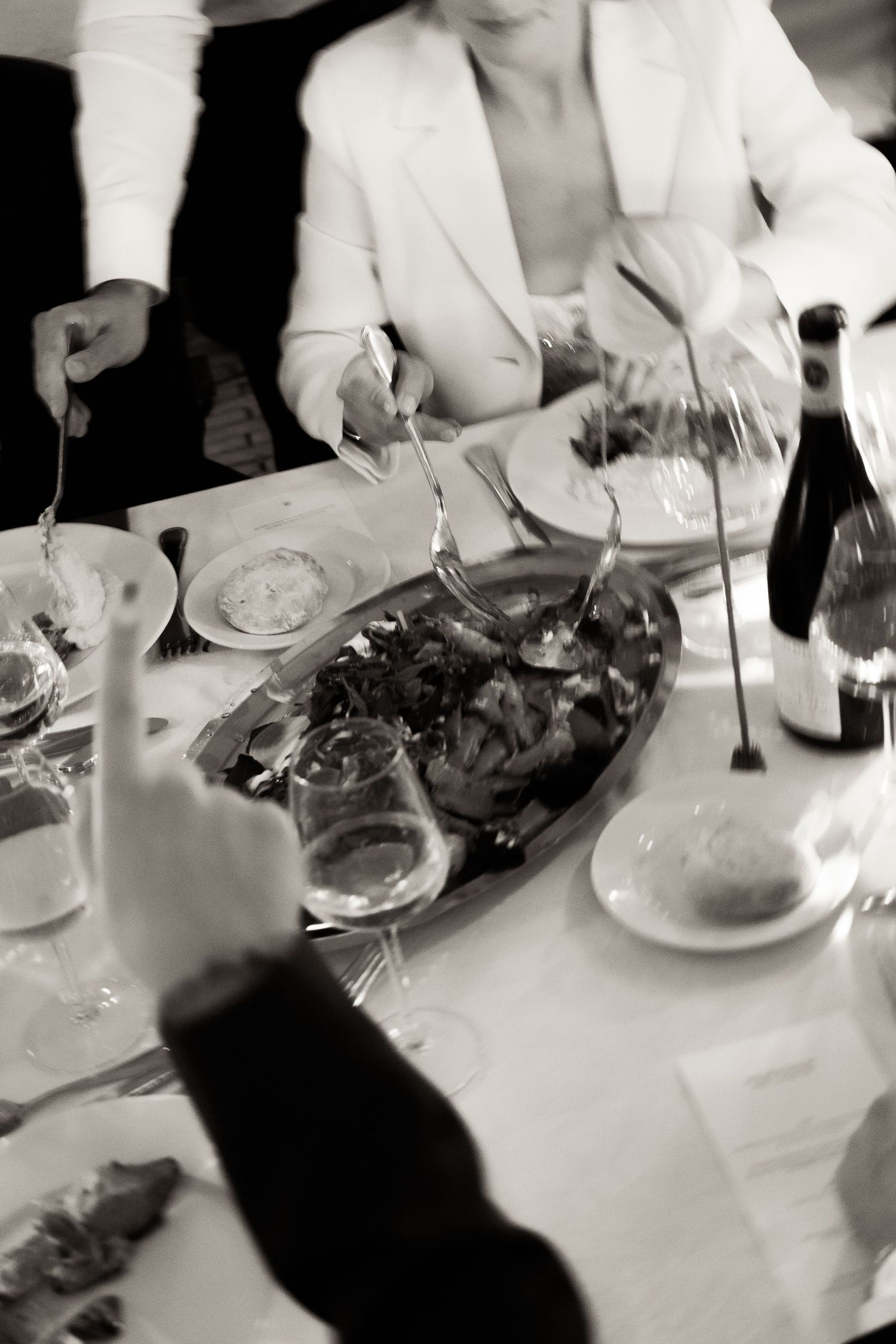 Black and white image of people dining at a formal restaurant table with plates of food and white tablecloth.
