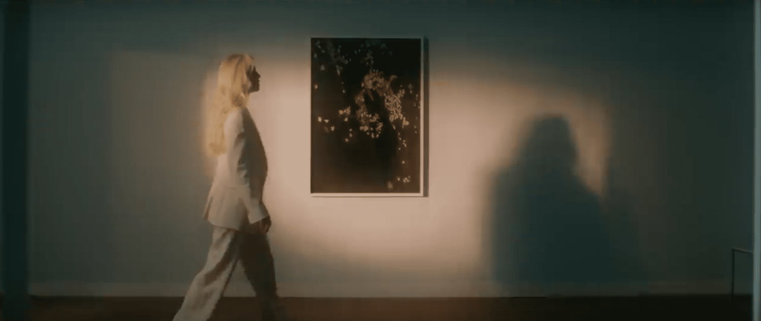 A blurry figure walks through a dimly lit gallery space with white walls displaying artwork.