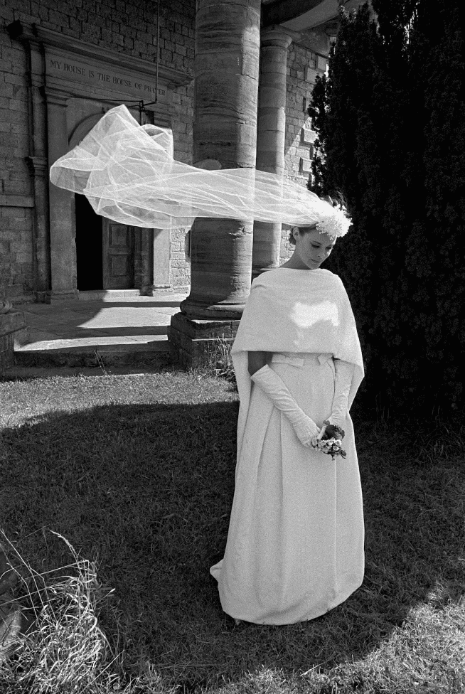 A ghostly figure in flowing white fabric stands among columns and grass in an artistic black and white photograph.