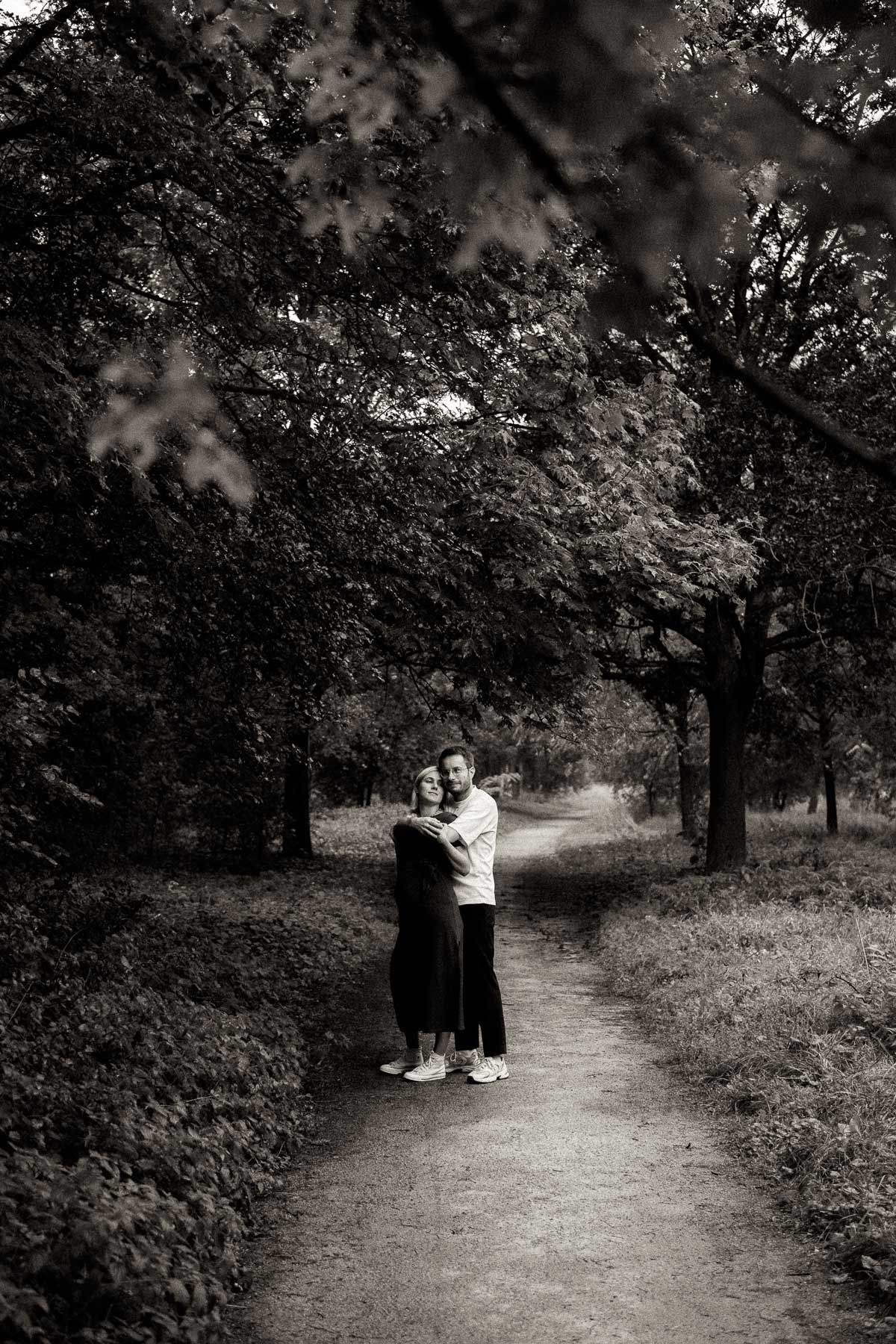 A couple walks together on a winding dirt path through a wooded area in black and white photography.