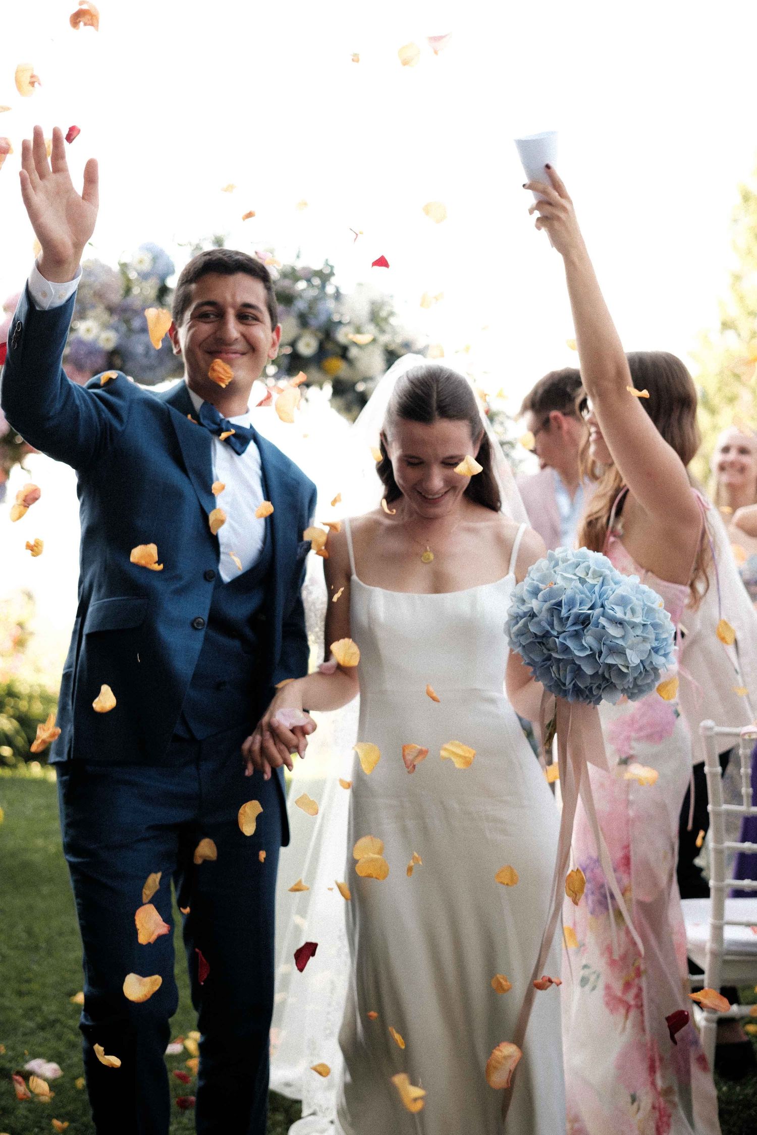 Wedding celebration with flower petals falling as couple walks down aisle while guests cheer on sunny outdoor day.