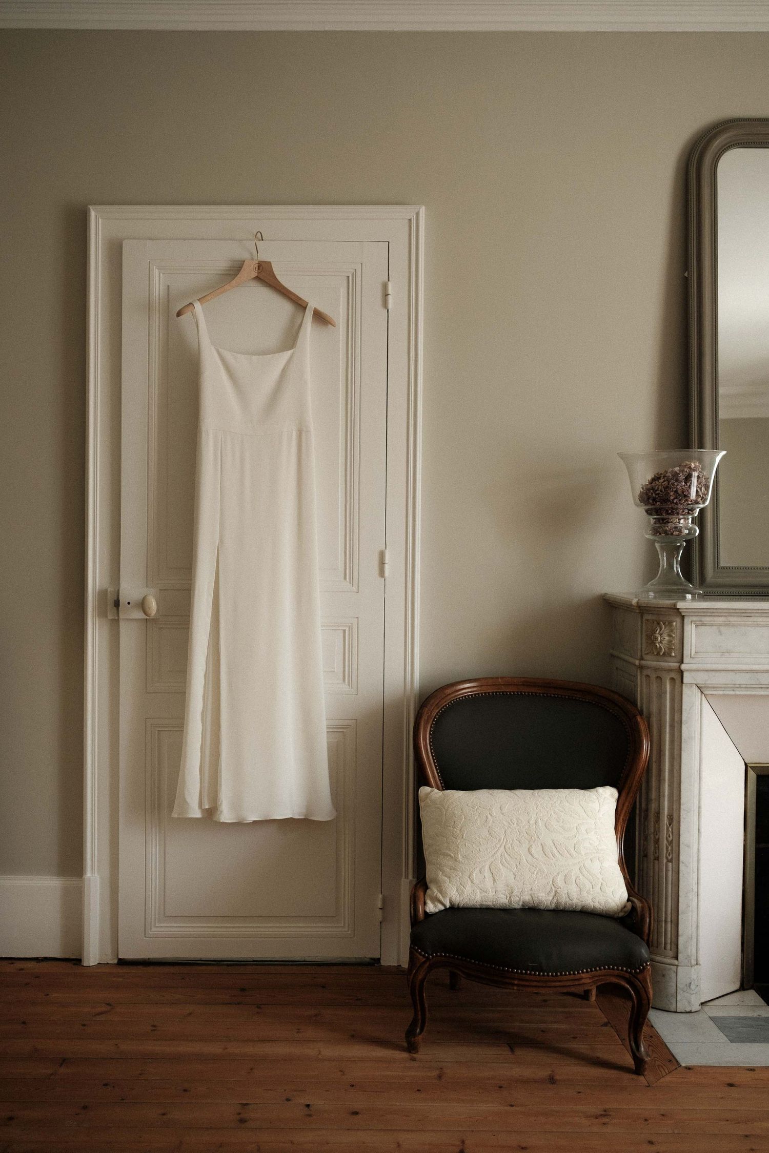A white dress hangs on a hanger against a cream wall with dark wood furniture and minimalist decor.