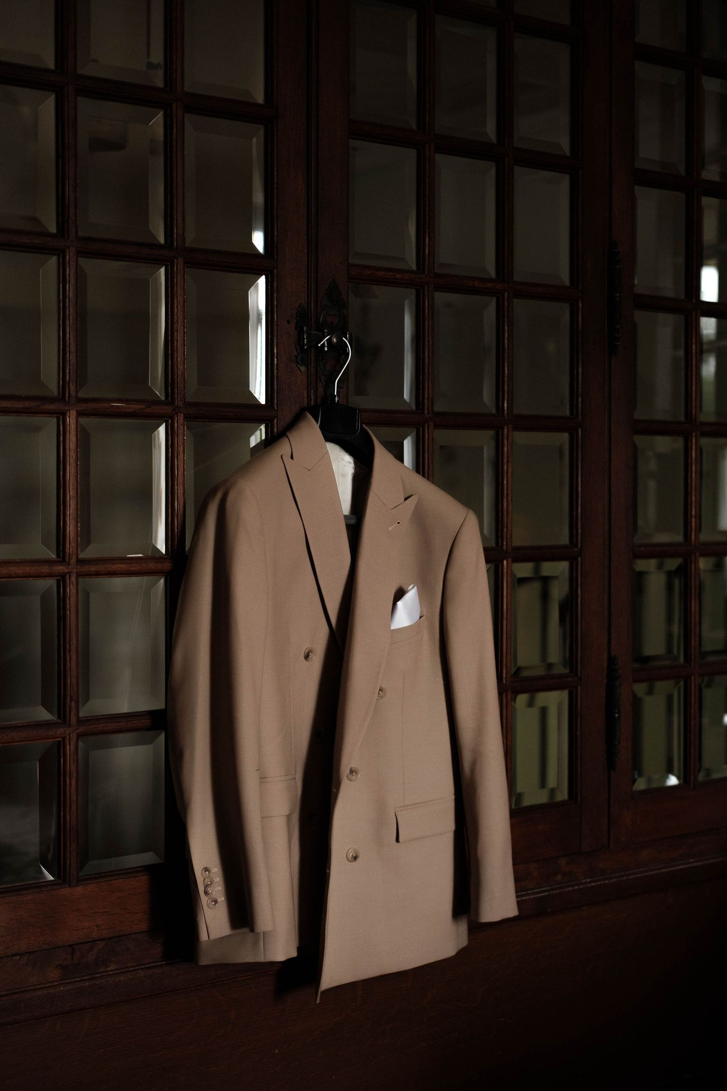 A tan blazer with white pocket square hangs on display against dark wooden paneled walls.