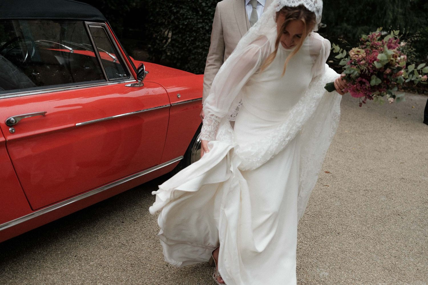 A flowing white wedding dress billows in the wind next to a vintage red car with pink flowers in the background.