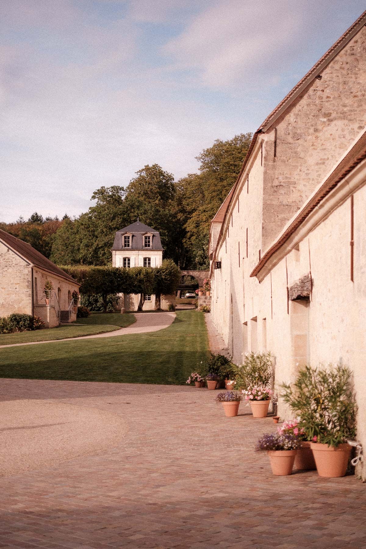 Stone buildings with terracotta pots line a courtyard path leading to an elegant mansion on a hill in warm evening light.