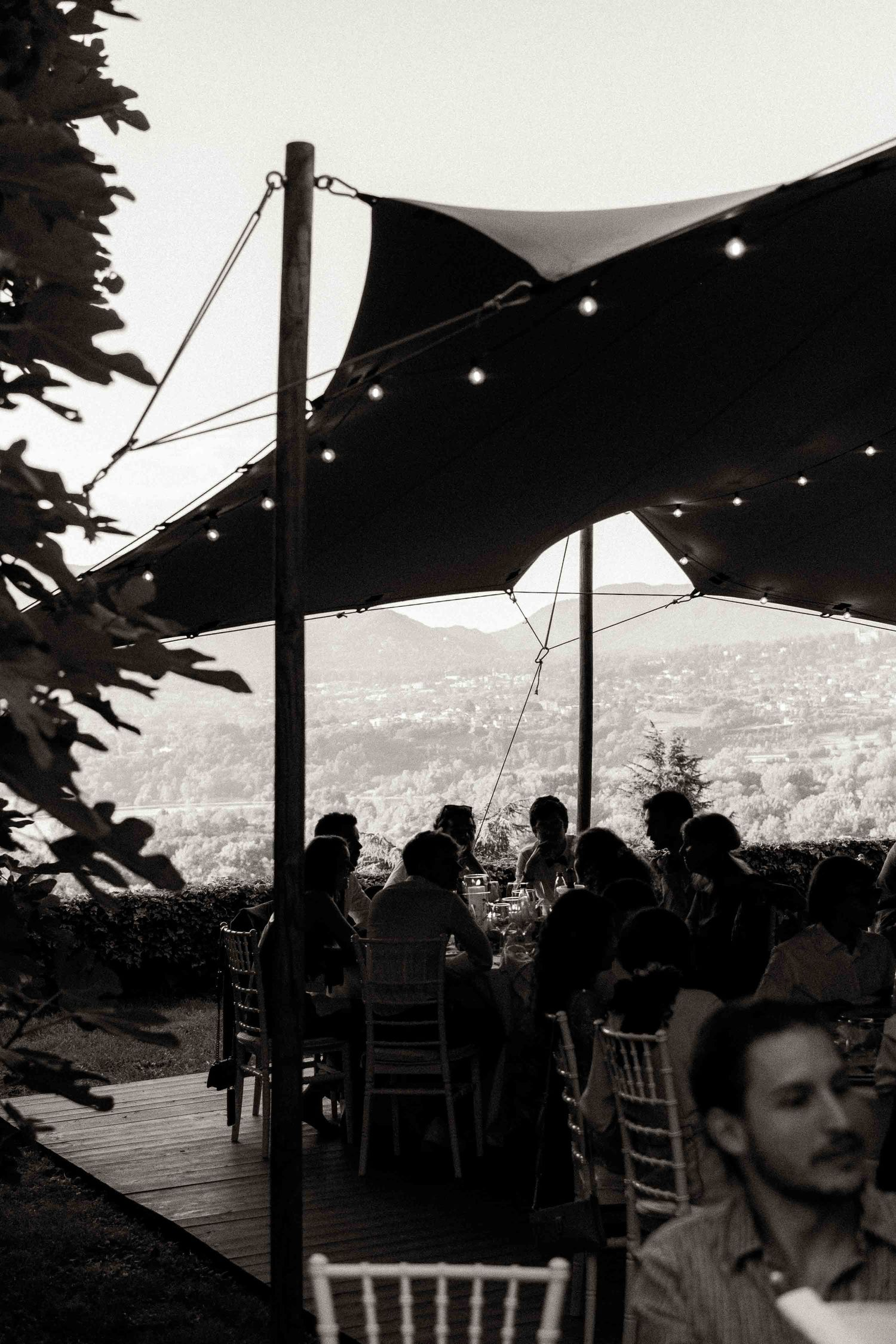 People gather under a dark canopy tent at an outdoor event overlooking a scenic mountain landscape in black and white.