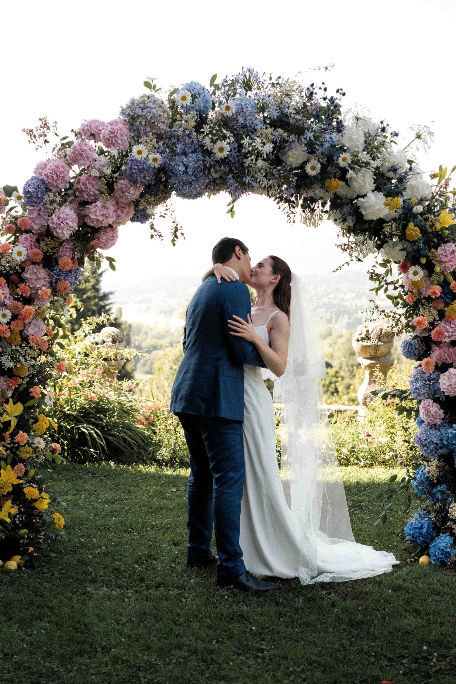 A couple shares a romantic kiss under a stunning floral arch with pink, blue and yellow blooms in an outdoor garden setting.