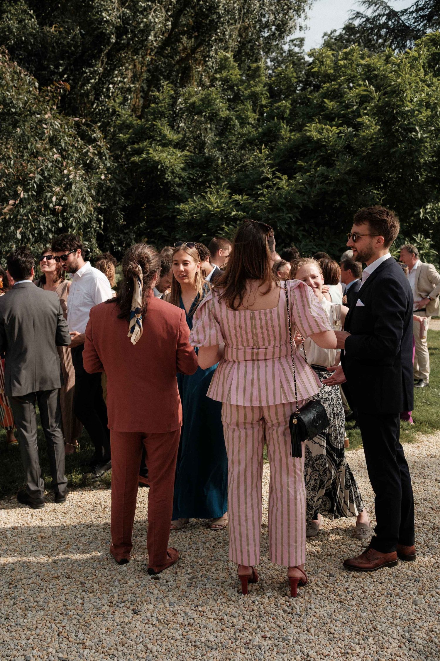 A group of wedding guests in formal attire stand together on a gravel path during an outdoor celebration.