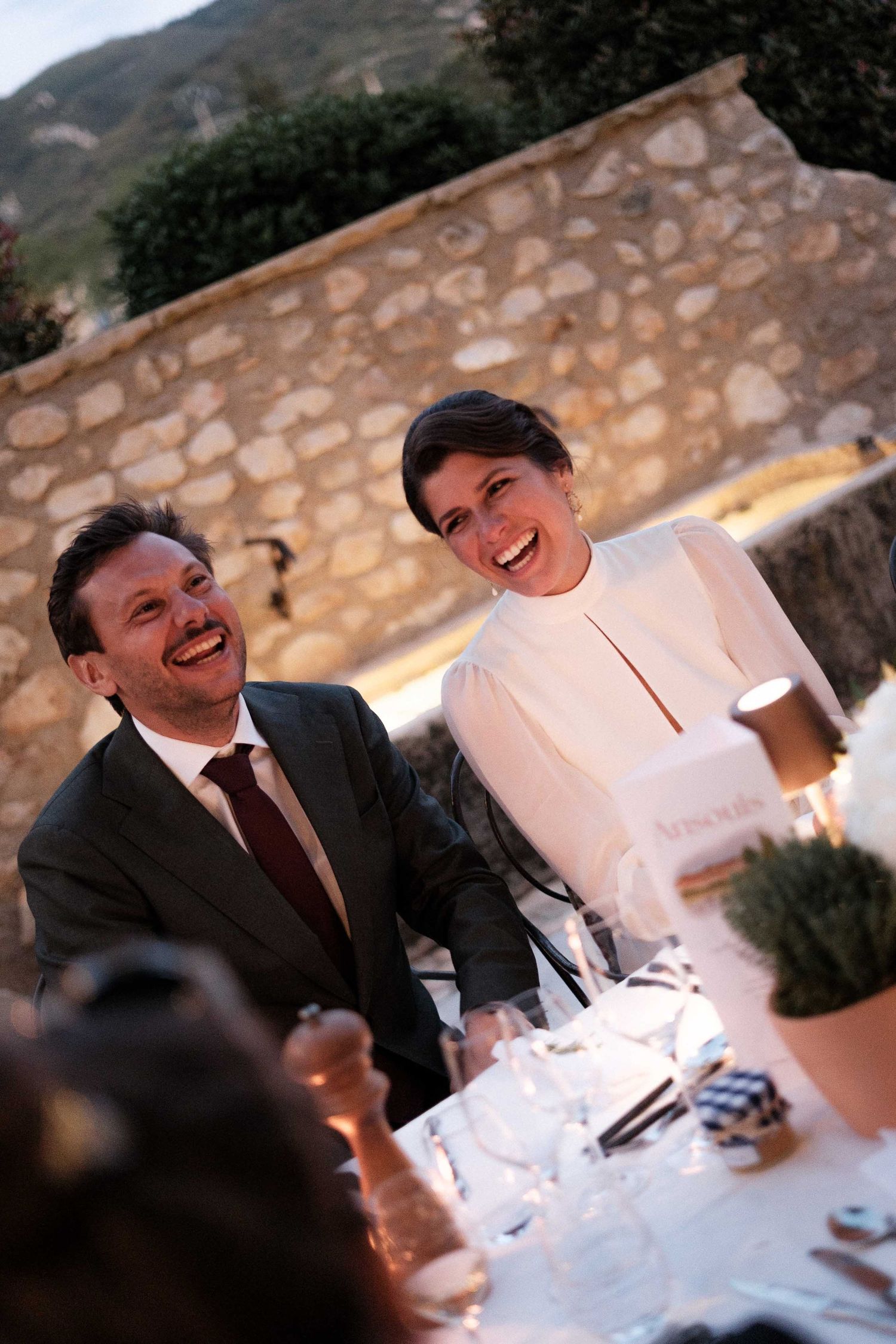 People share joyful laughter during an outdoor dinner gathering at sunset against a stone wall backdrop.