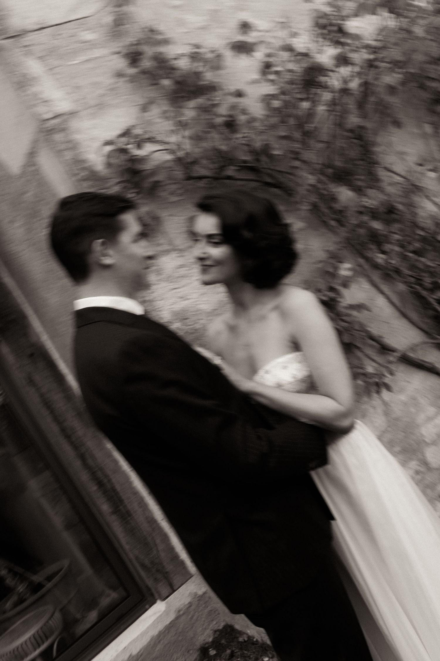 A black and white vintage-style wedding photograph showing a romantic moment between a couple in formal attire.