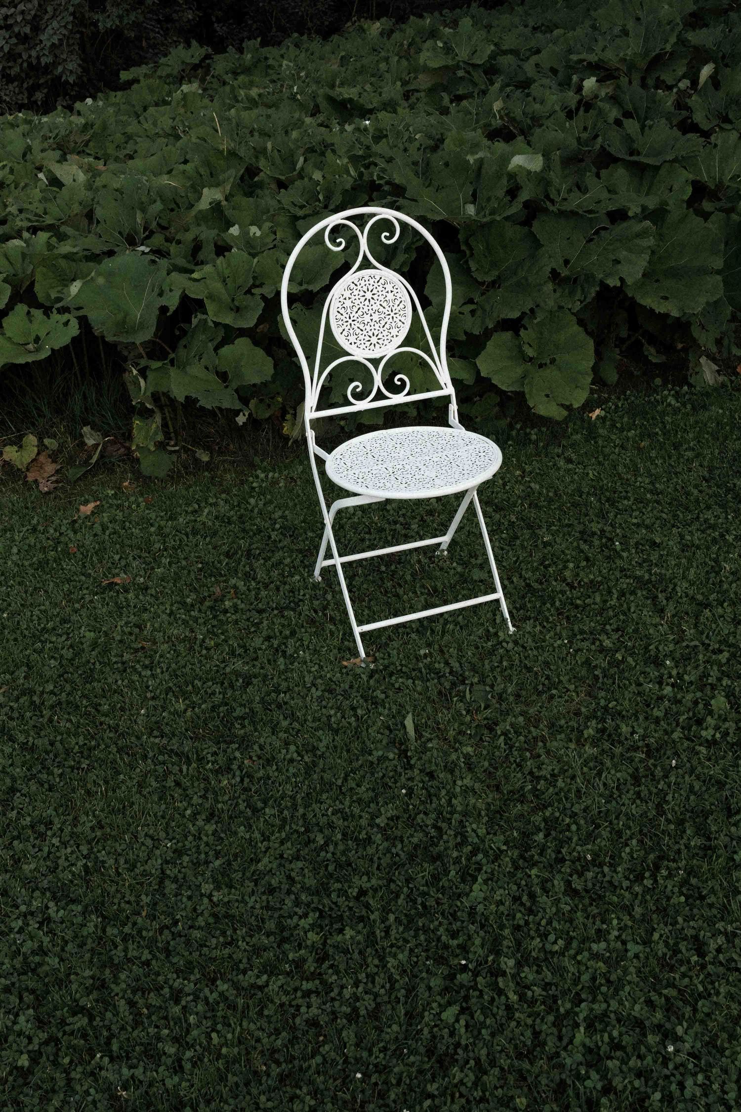 A decorative white wrought iron garden bench sits empty on green grass with trees in the background.