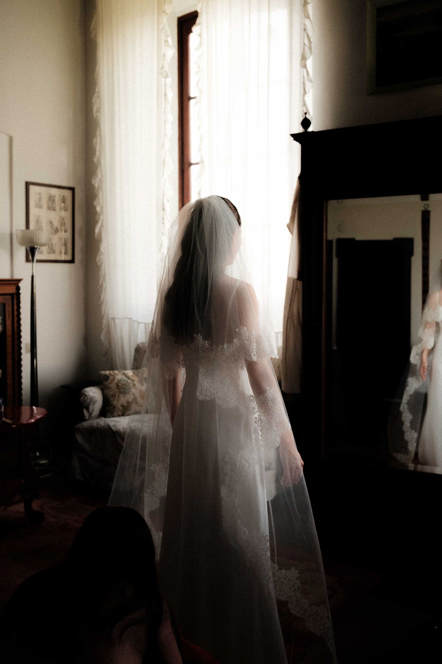 A bride in a white wedding dress and veil stands silhouetted against a bright window in a dimly lit room.