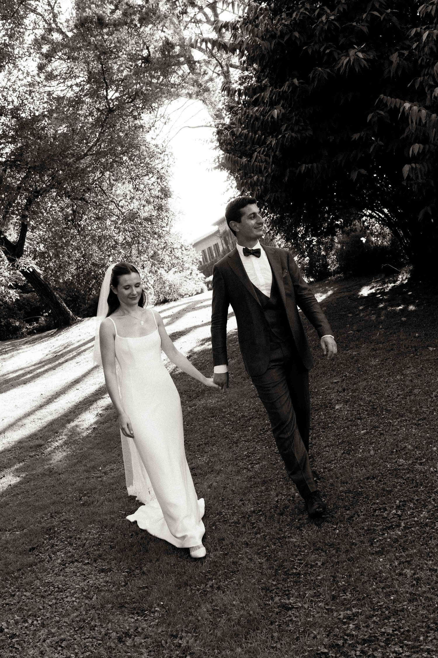 A couple walks hand in hand along a tree-lined path in black and white wedding attire.