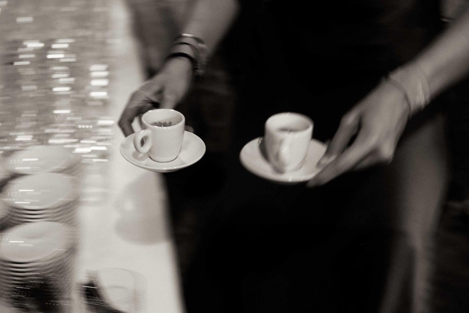 Hands serving espresso cups on white saucers in a black and white artistic cafe setting.