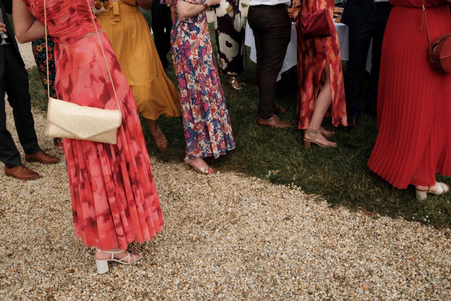 A group of people in red and floral dresses gather outdoors on a gravel path during a social event.