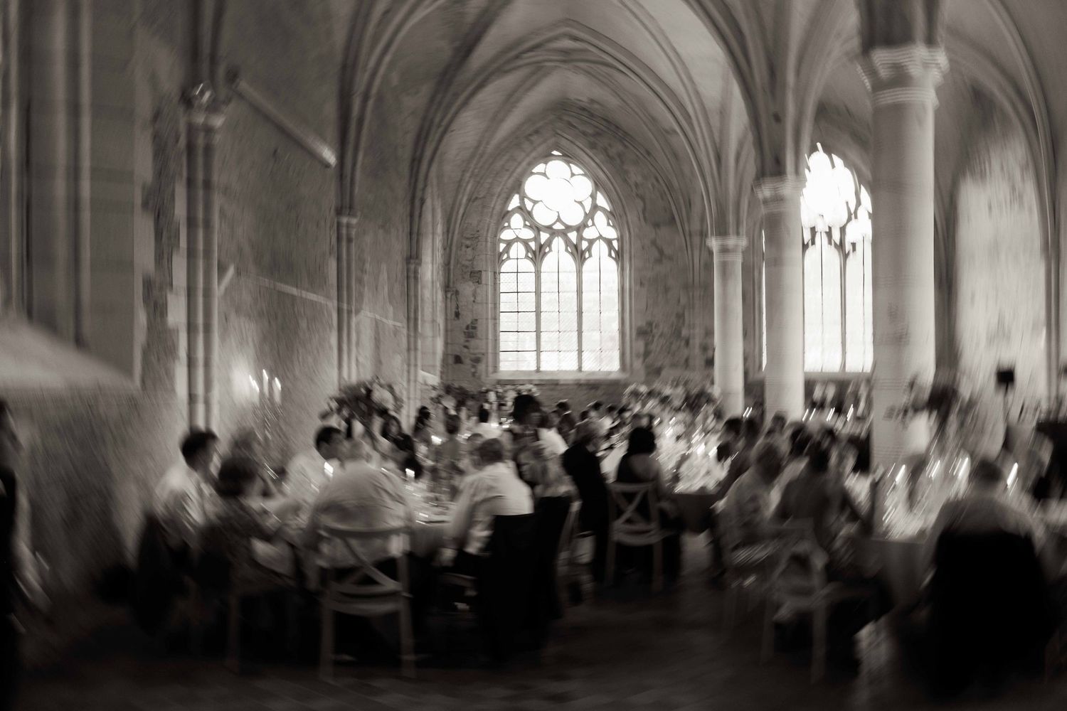 A blurred black and white photograph shows people gathered in a Gothic cathedral dining hall with vaulted ceilings and windows.