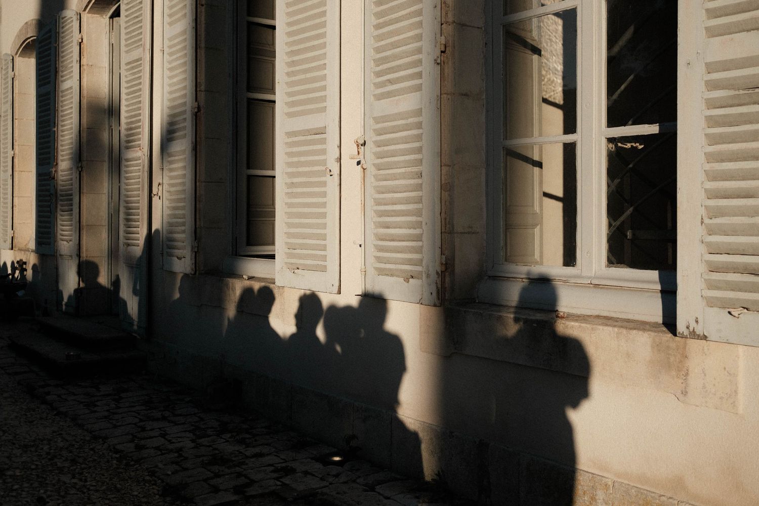 Silhouettes of people cast shadows on a white wall with shuttered windows in an architectural passageway.
