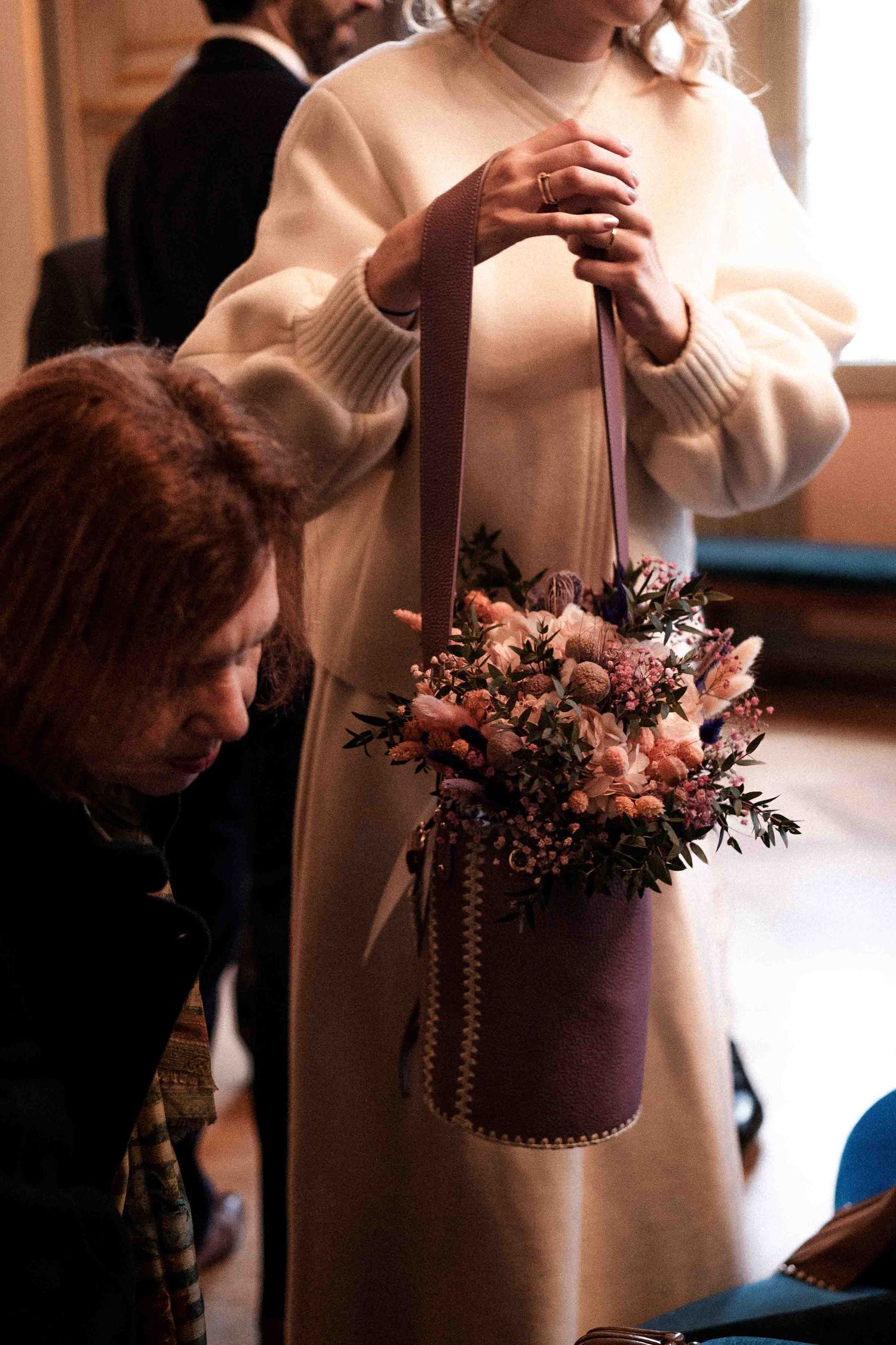 A person in a cream-colored sweater holds a delicate pink and white floral arrangement in warm lighting.