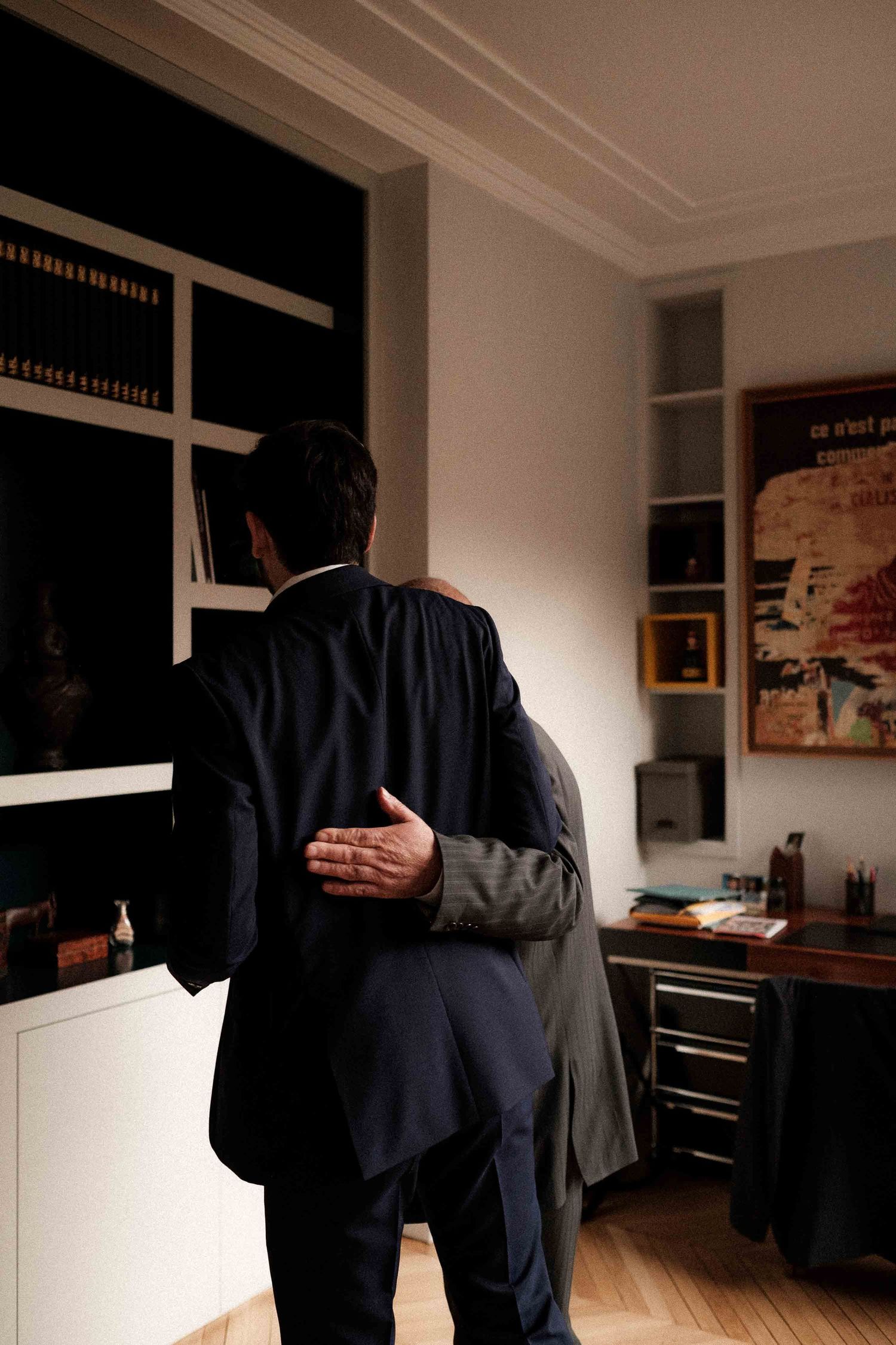 Dark silhouettes of figures in formal attire stand in a dimly lit room with white walls and artwork.