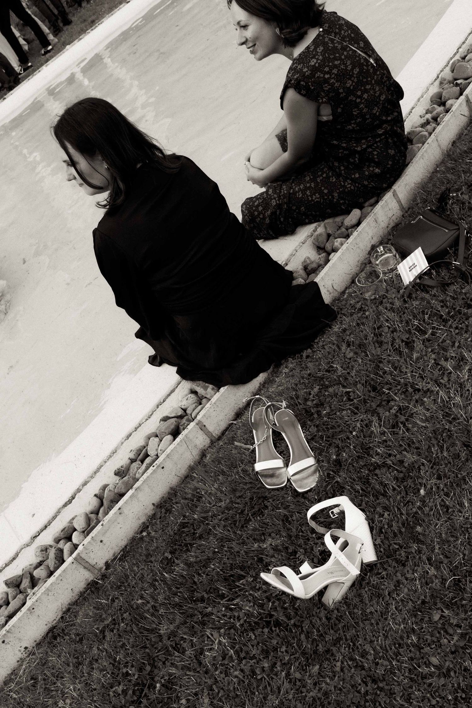 Two people sitting on a curb with white high-heeled sandals placed on grass beside them in black and white.