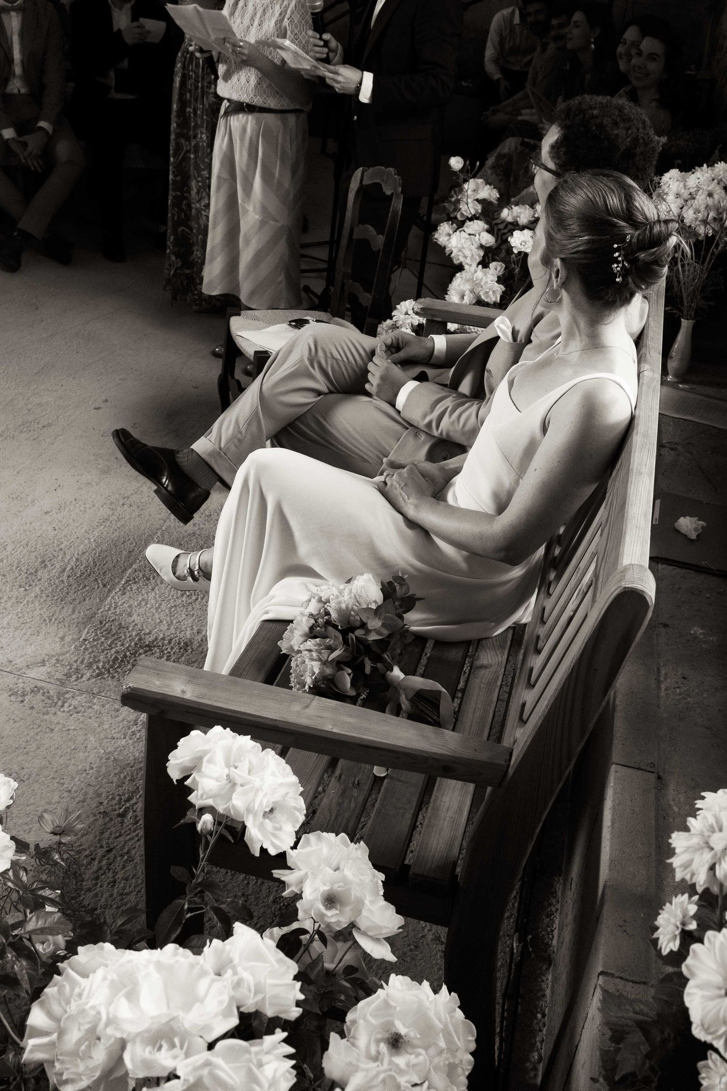 A black and white photo shows someone sitting in a chair beside white flowers at an indoor event.