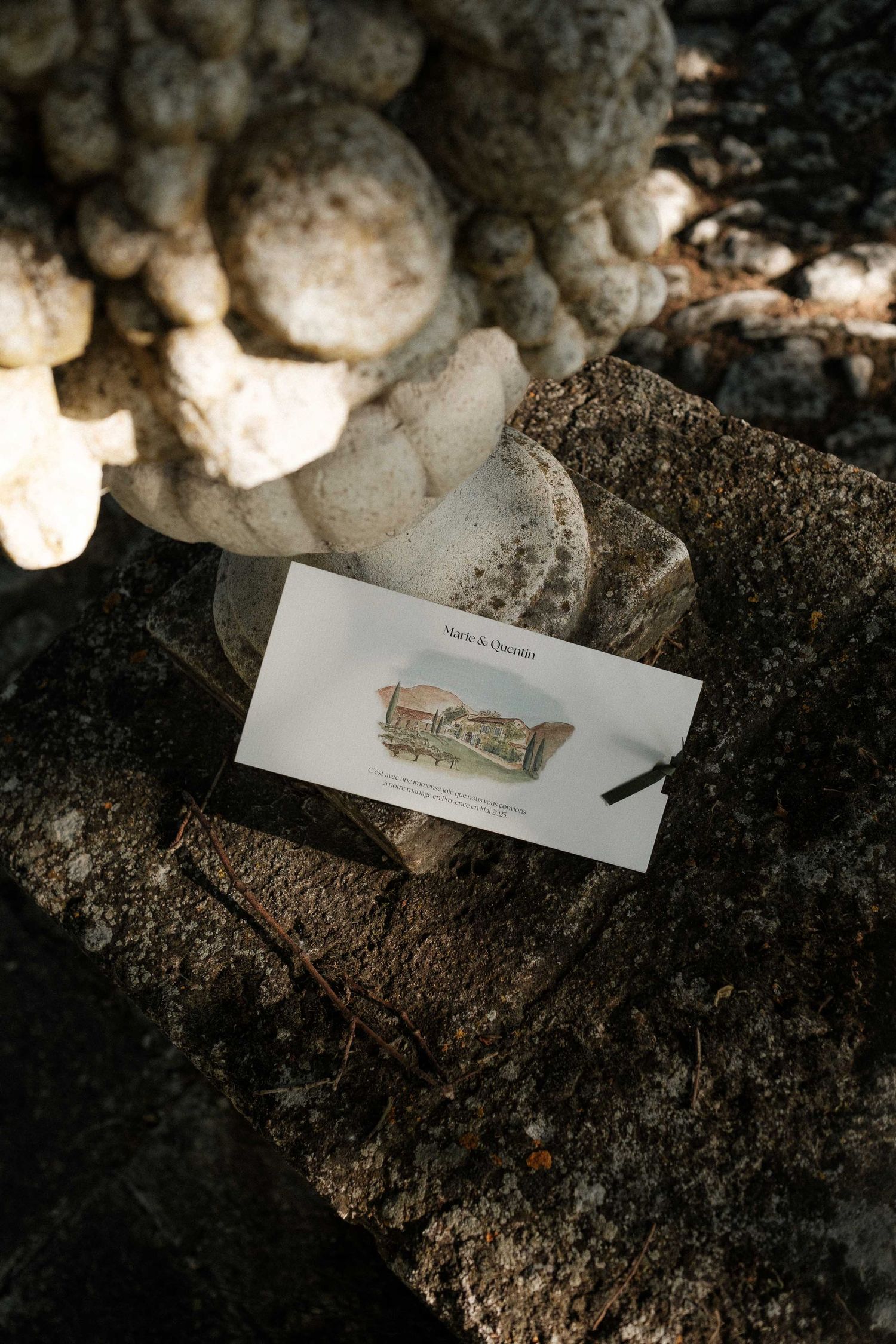 A small watercolor sketch of a building lies on dark soil next to white mushrooms in natural lighting.